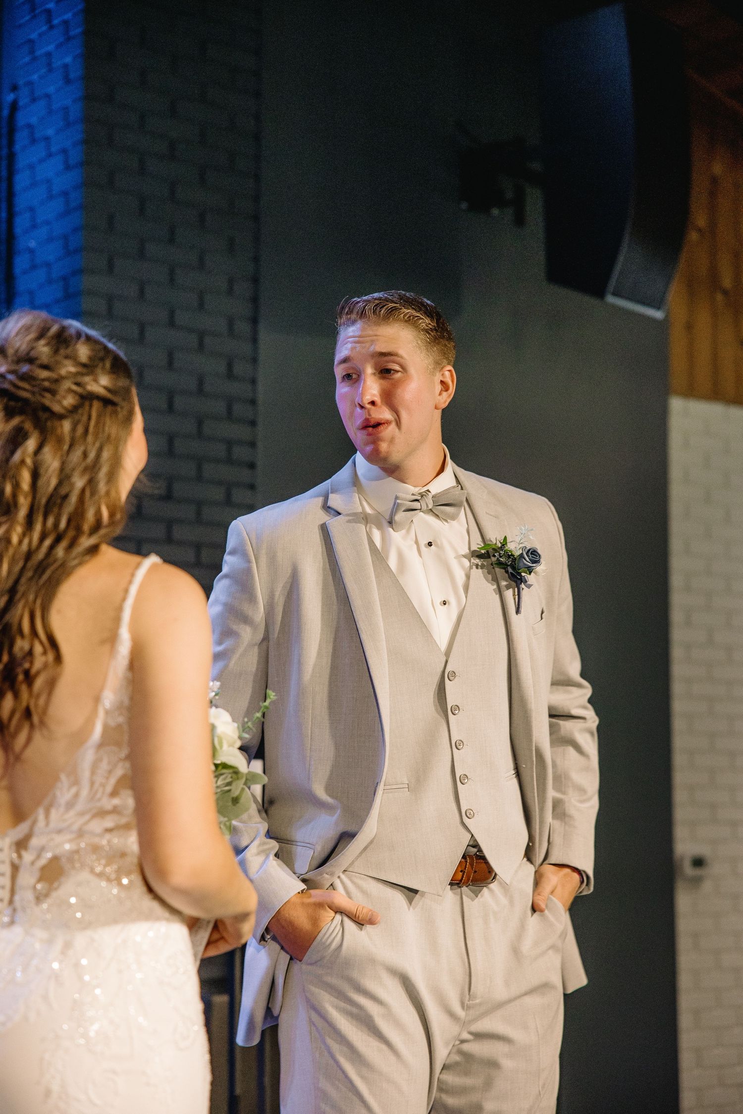 Groom in light gray suit with bow tie smiling at bride during wedding ceremony indoors.
