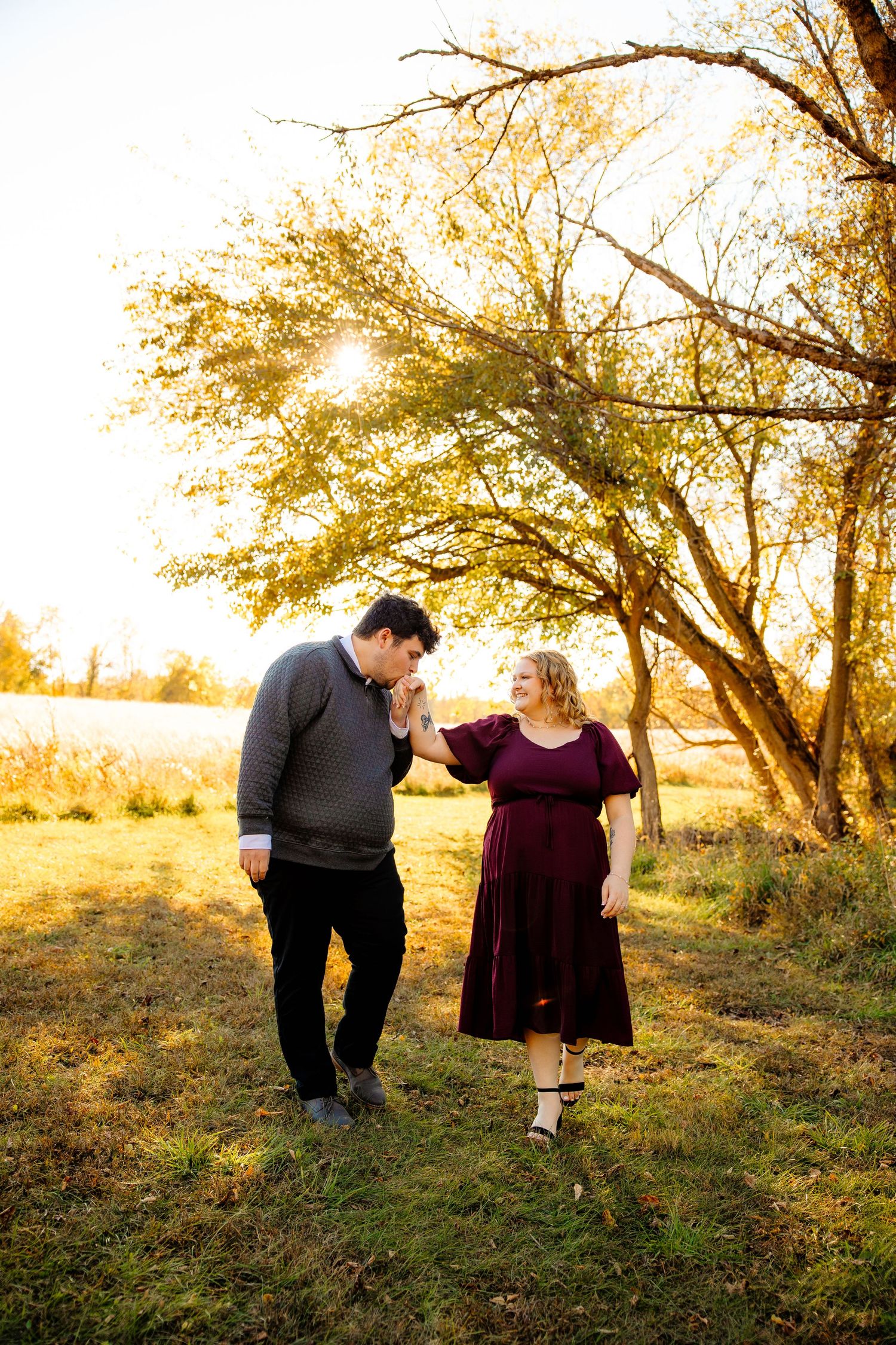 A couple shares a romantic moment at golden hour, man kissing woman's hand in a sunlit autumn field.