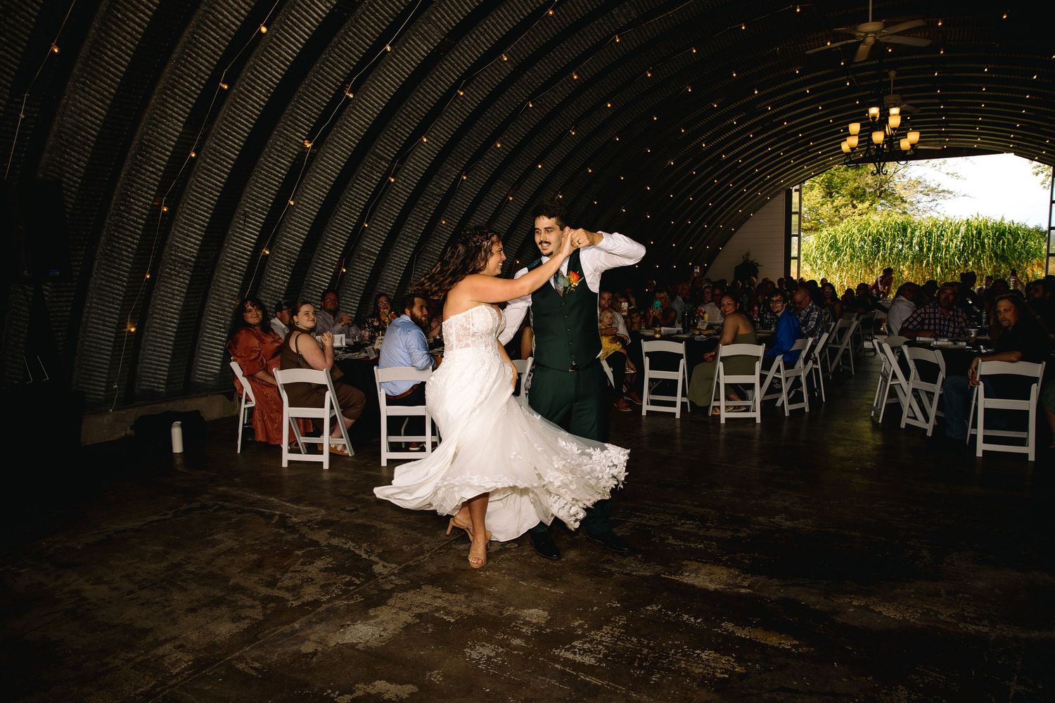 Bride and groom share first dance in rustic barn venue with guests seated around them.
