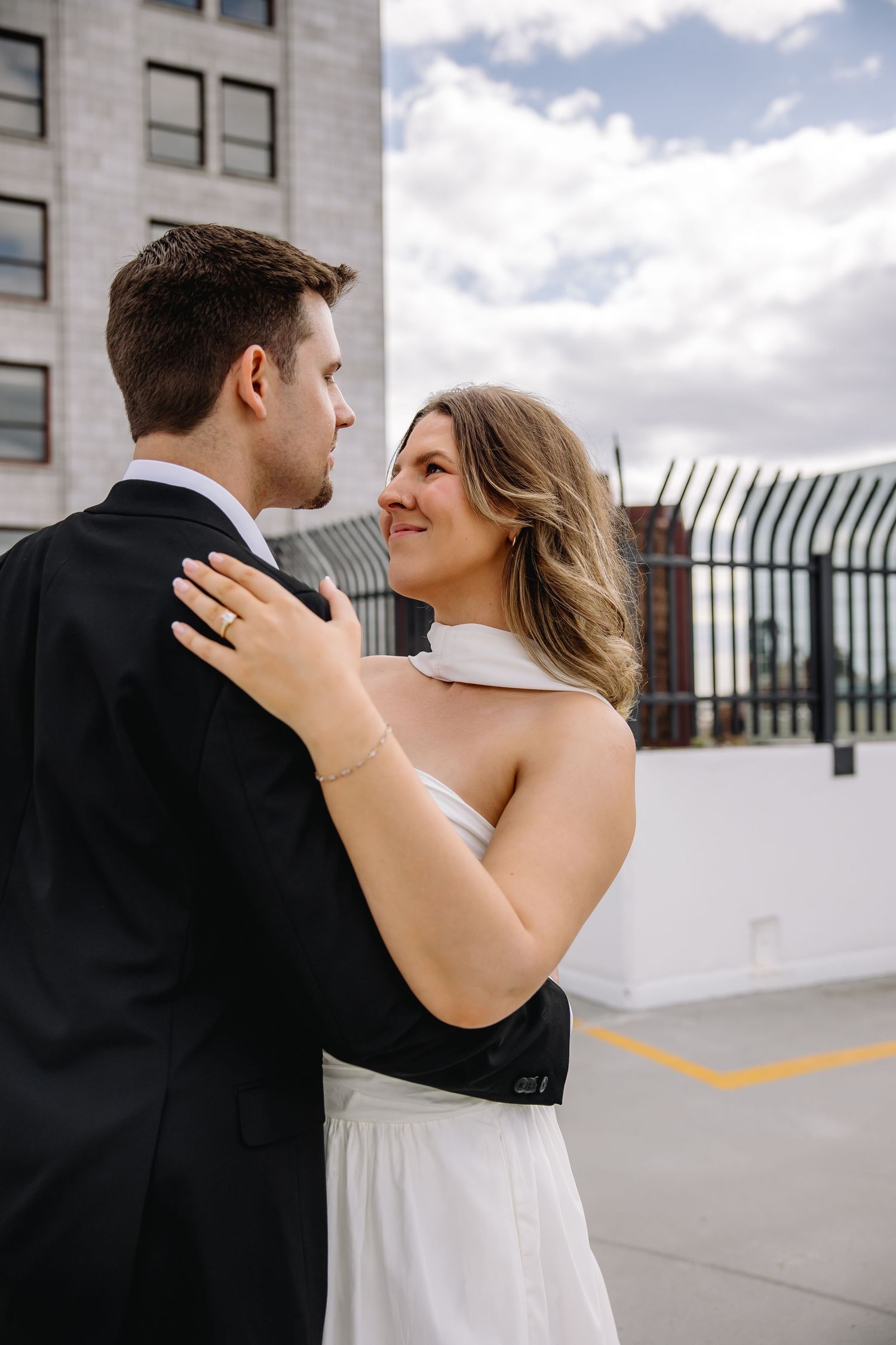 A bride in a white halter gown embraces her groom in a black suit on a rooftop, smiling lovingly on their wedding day.