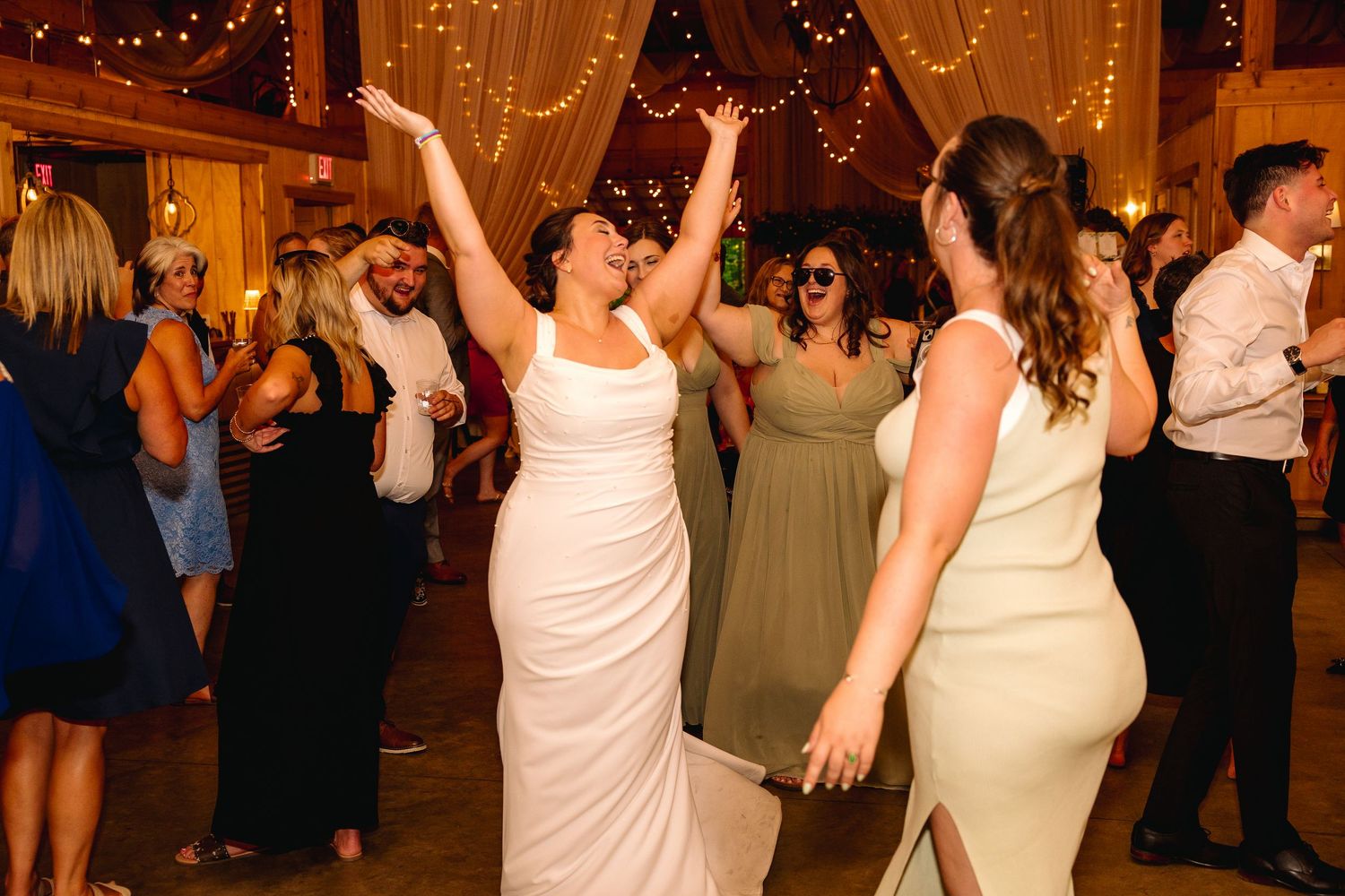 Two brides in white gowns dance joyfully with guests at a warmly lit wedding reception venue.