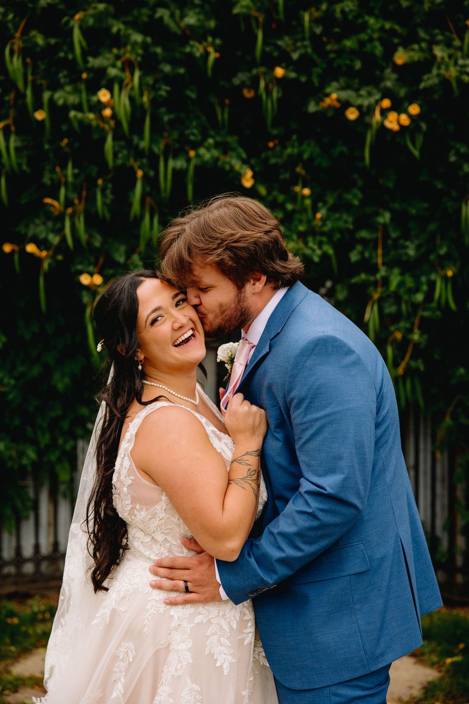 Bride in lace gown laughs joyfully as groom in blue suit kisses her cheek in a lush garden setting.