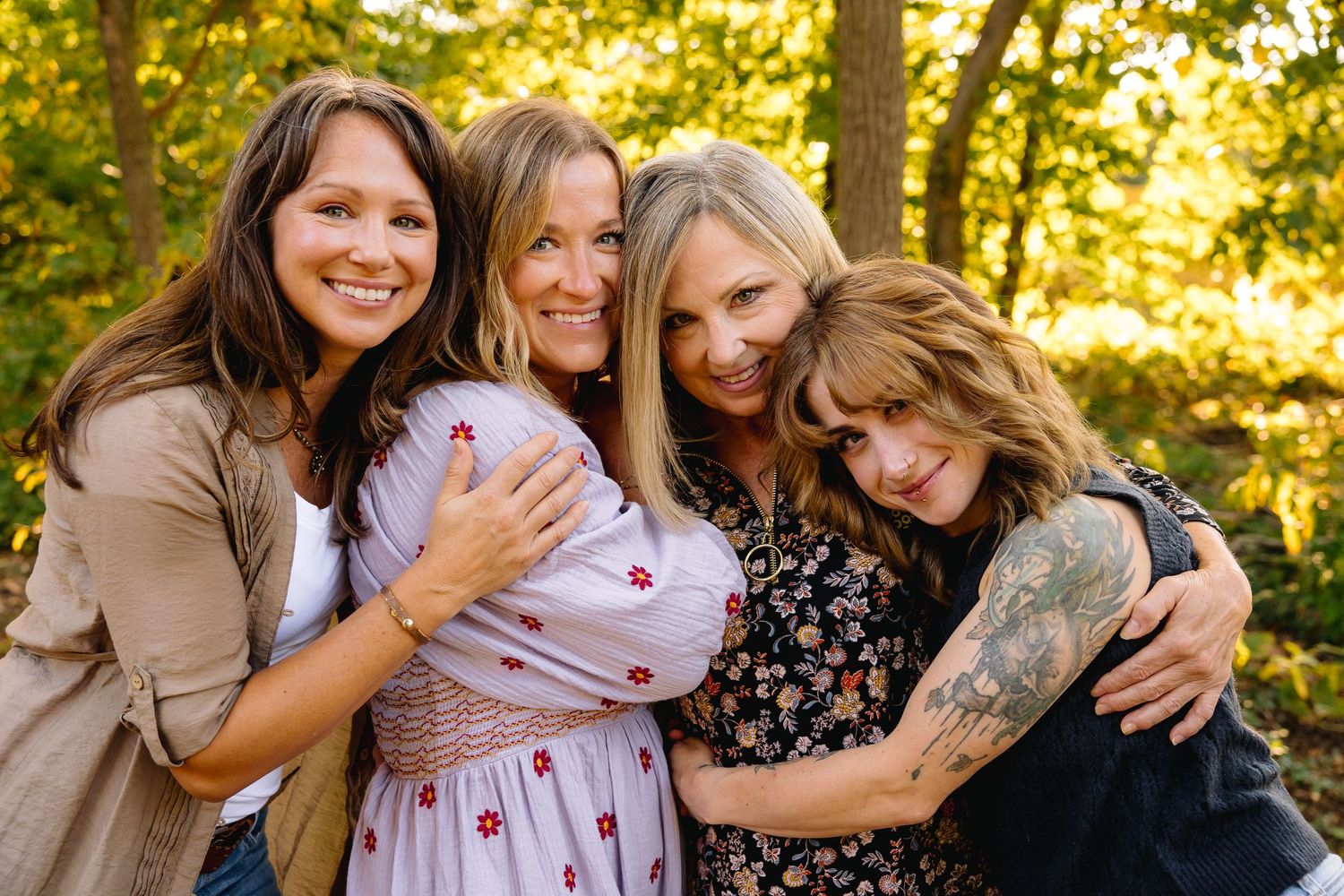 Four smiling women hugging outdoors among lush green trees, posing together for a warm group portrait.