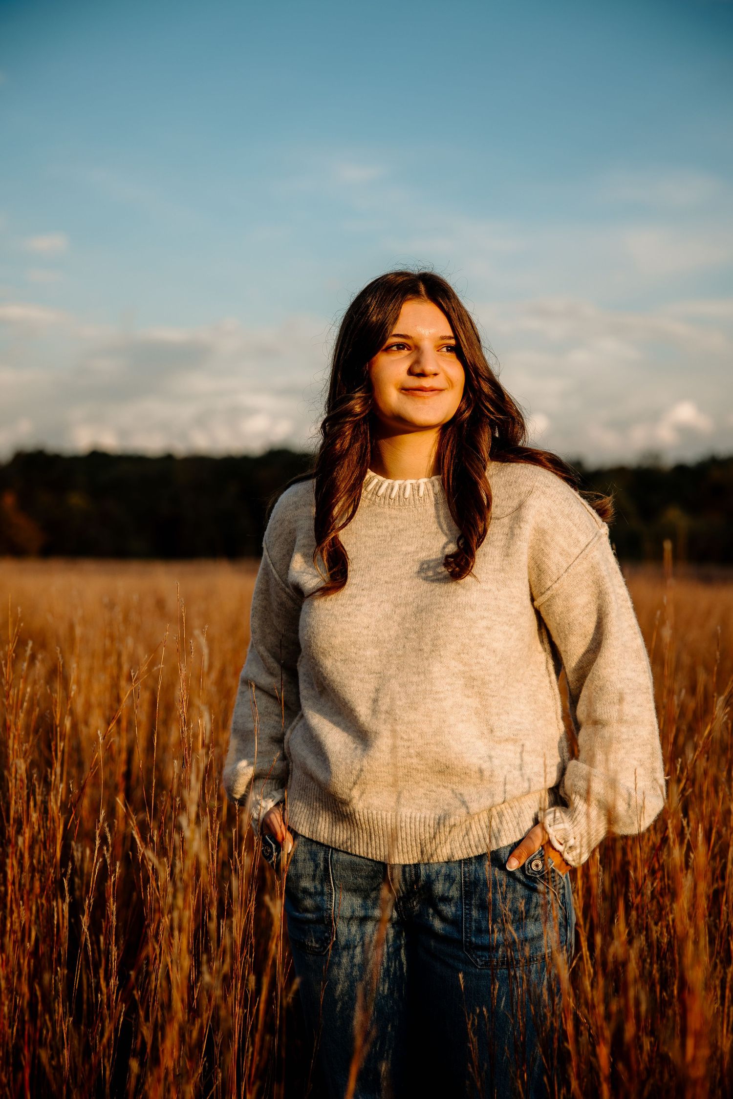 Young woman in gray sweater and jeans stands smiling in golden field at sunset with blue sky background.