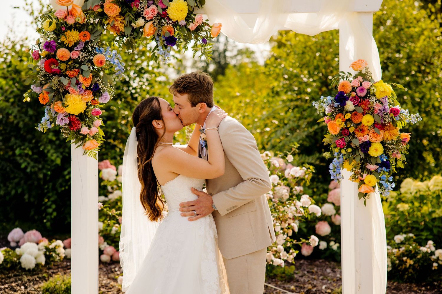 Bride and groom share first kiss under colorful floral arch with vibrant blooms at outdoor wedding ceremony.