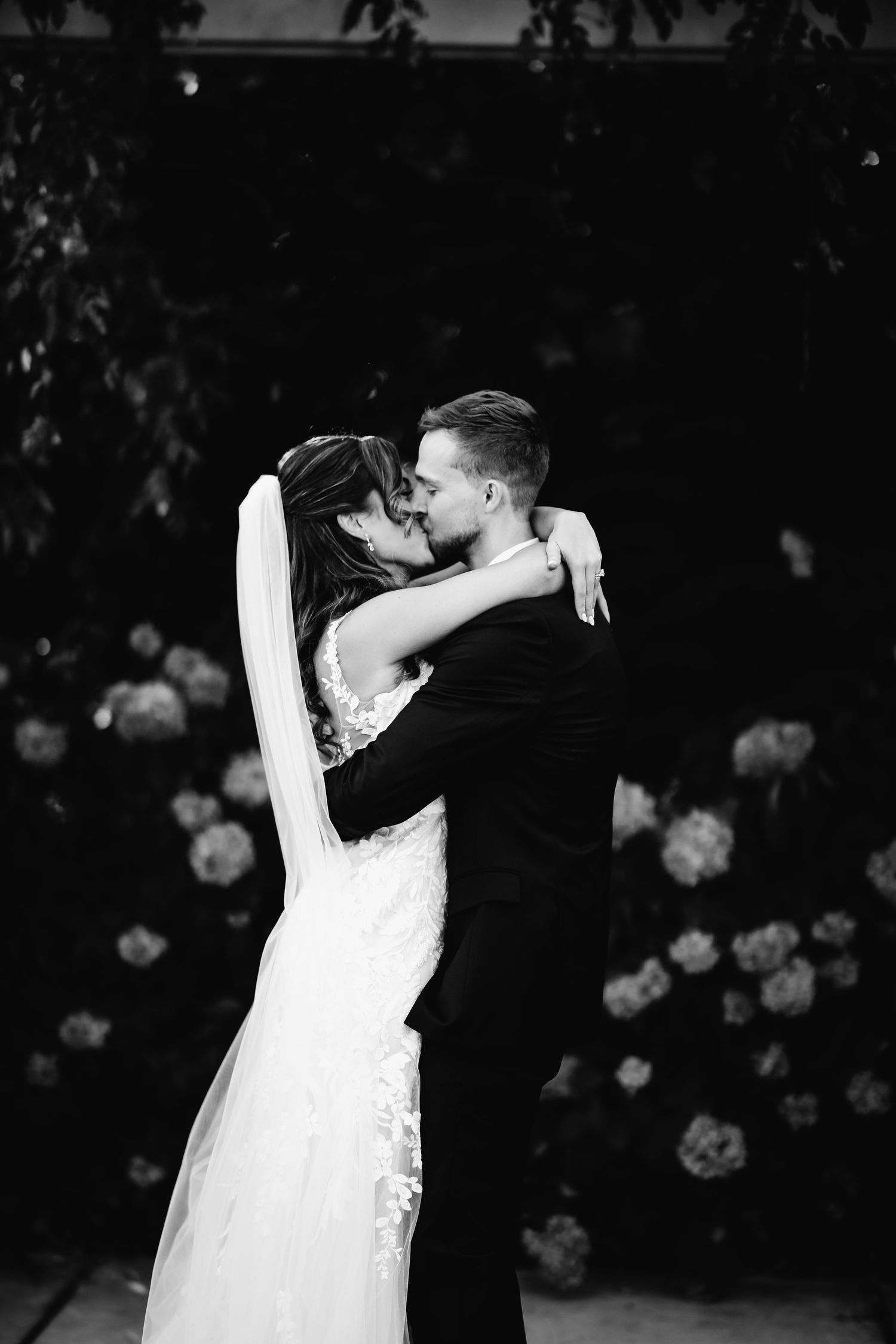 Black and white photo of a bride and groom sharing a romantic kiss at their wedding ceremony with floral backdrop.