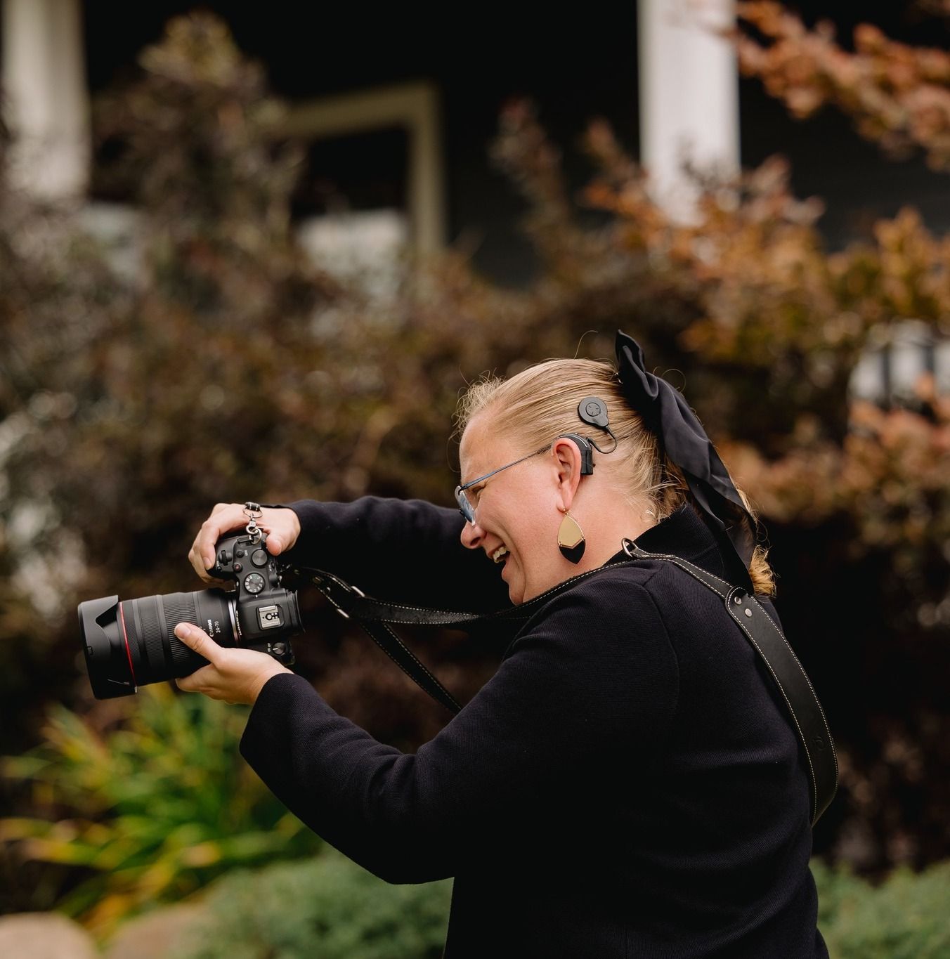 Smiling female photographer in black outfit laughing while holding DSLR camera outdoors in garden setting.