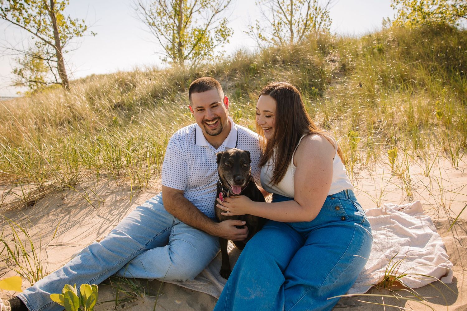 A happy couple sits on sandy dunes with their black dog, smiling in warm golden sunlight surrounded by green grass.