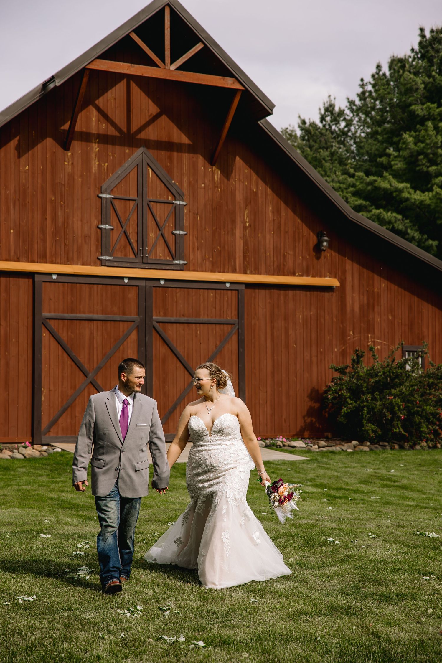 Bride and groom walk hand-in-hand on green grass in front of a rustic red barn on their wedding day.