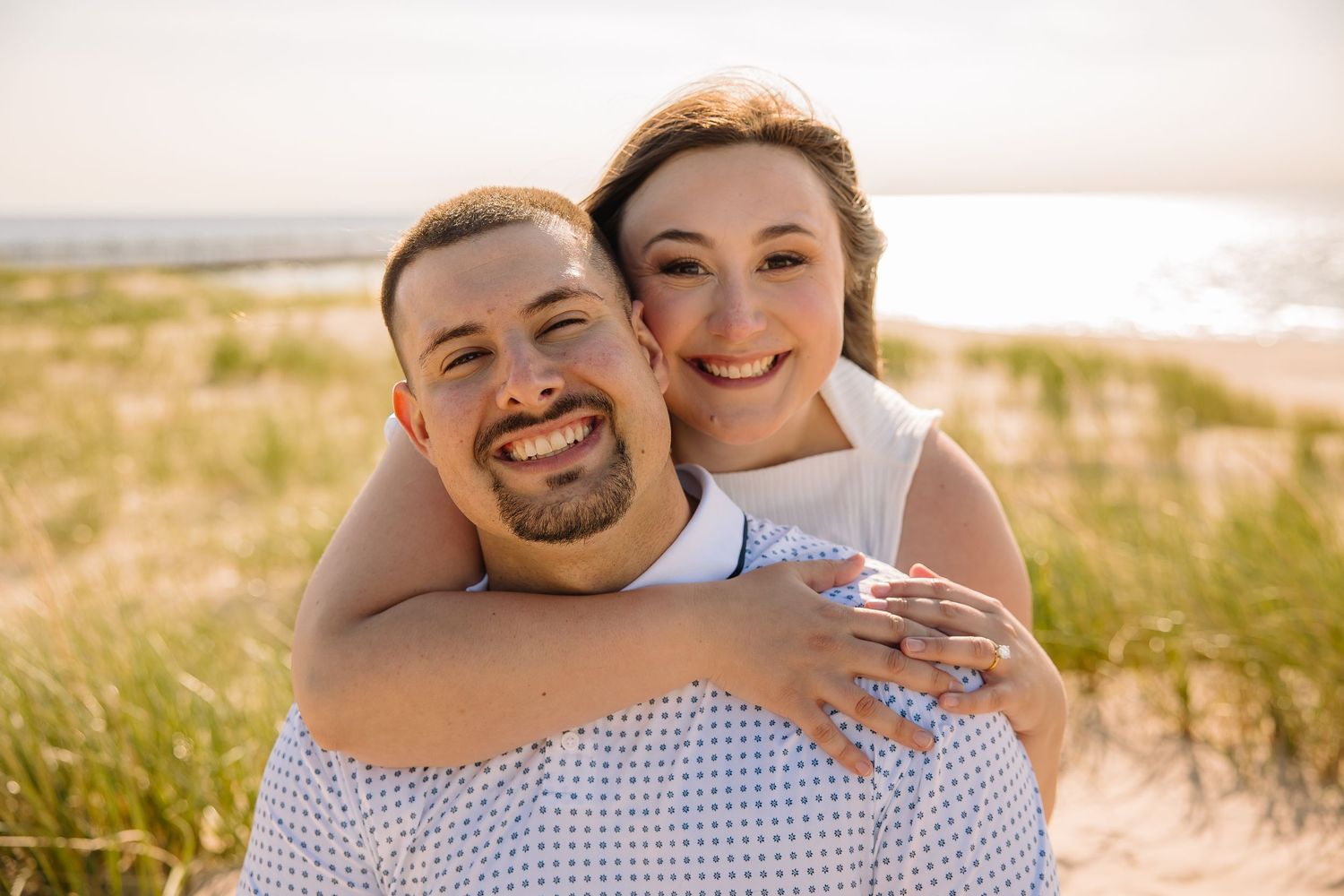 Happy couple embracing on a sandy beach, smiling warmly with ocean and dune grass in the background.