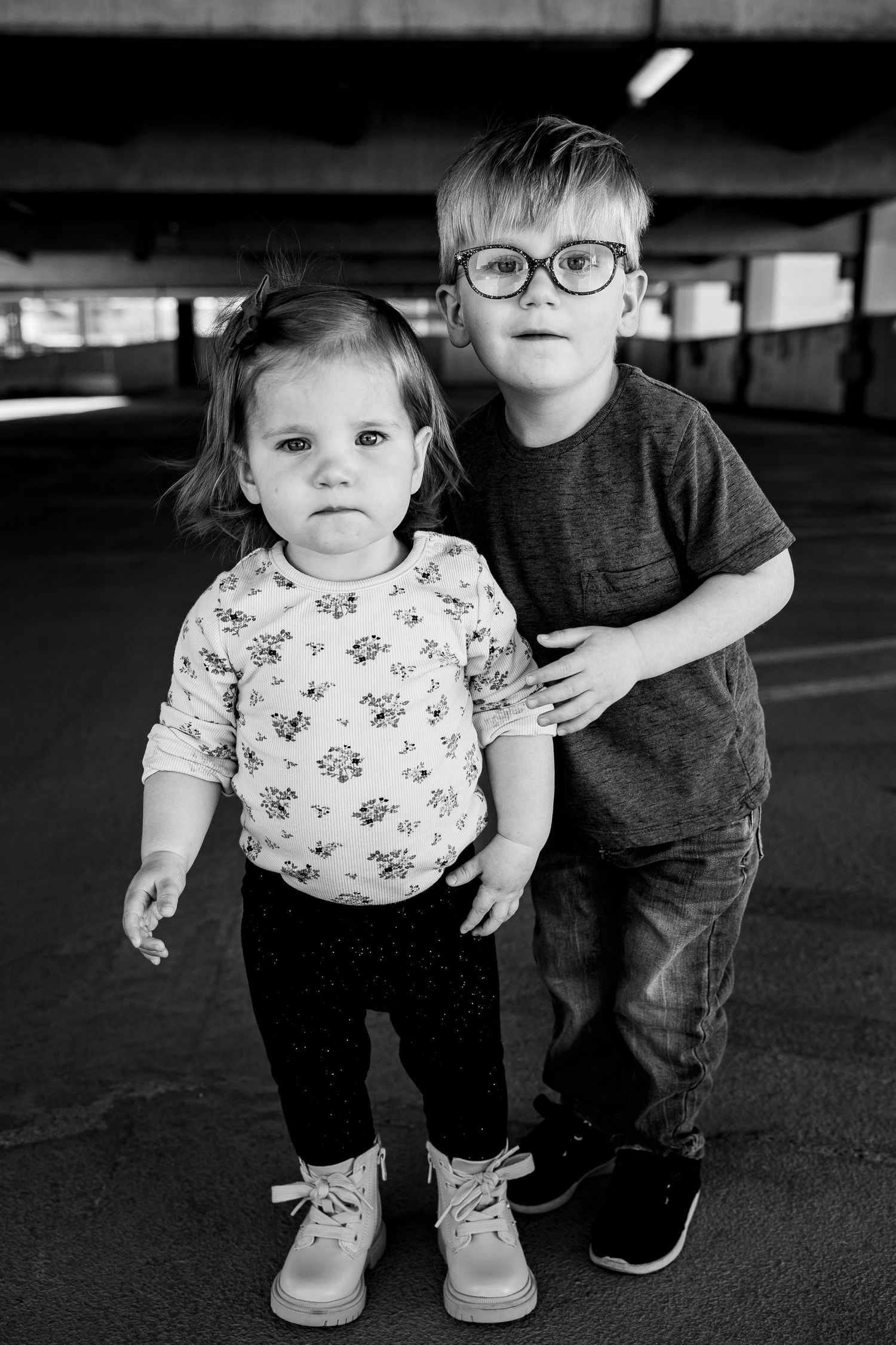 Black and white photo of two young siblings, a toddler girl and older boy wearing glasses, posing in a parking garage.