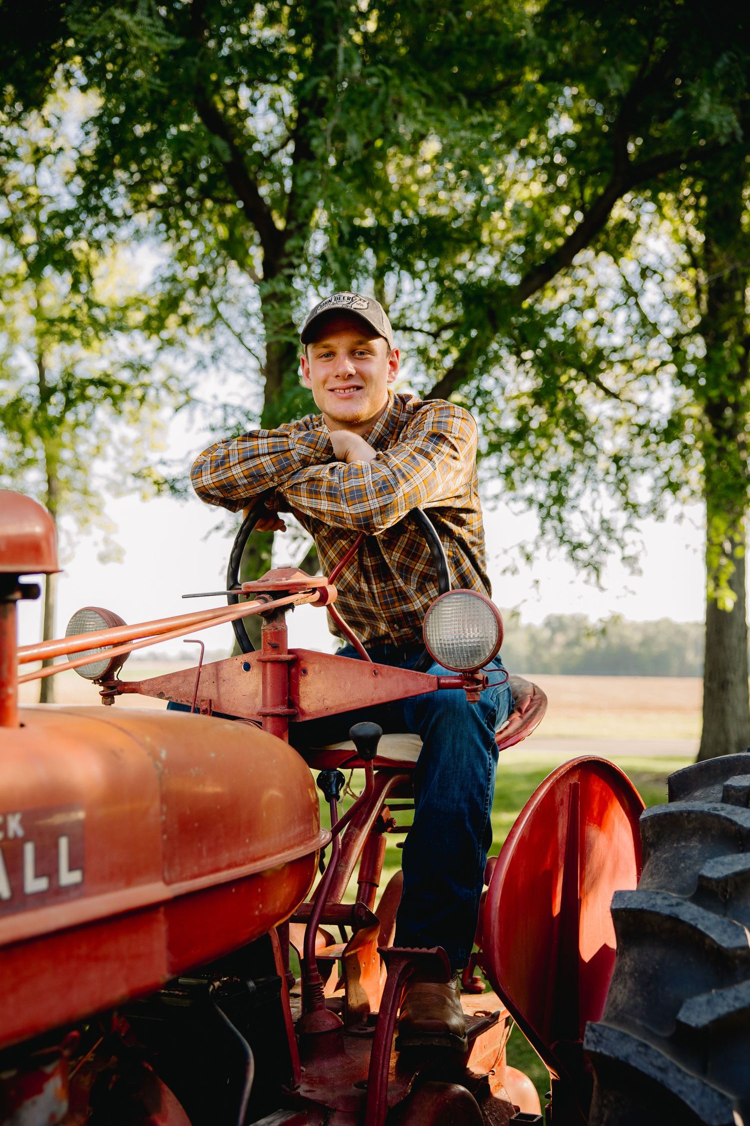Young man in plaid shirt and cap sitting on a vintage red Farmall tractor outdoors near trees.
