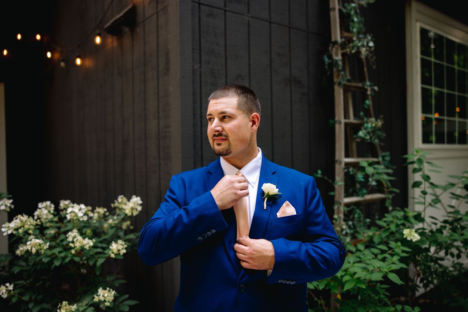 Groom in royal blue suit adjusting tie with white boutonniere, posing outdoors near rustic barn with string lights.