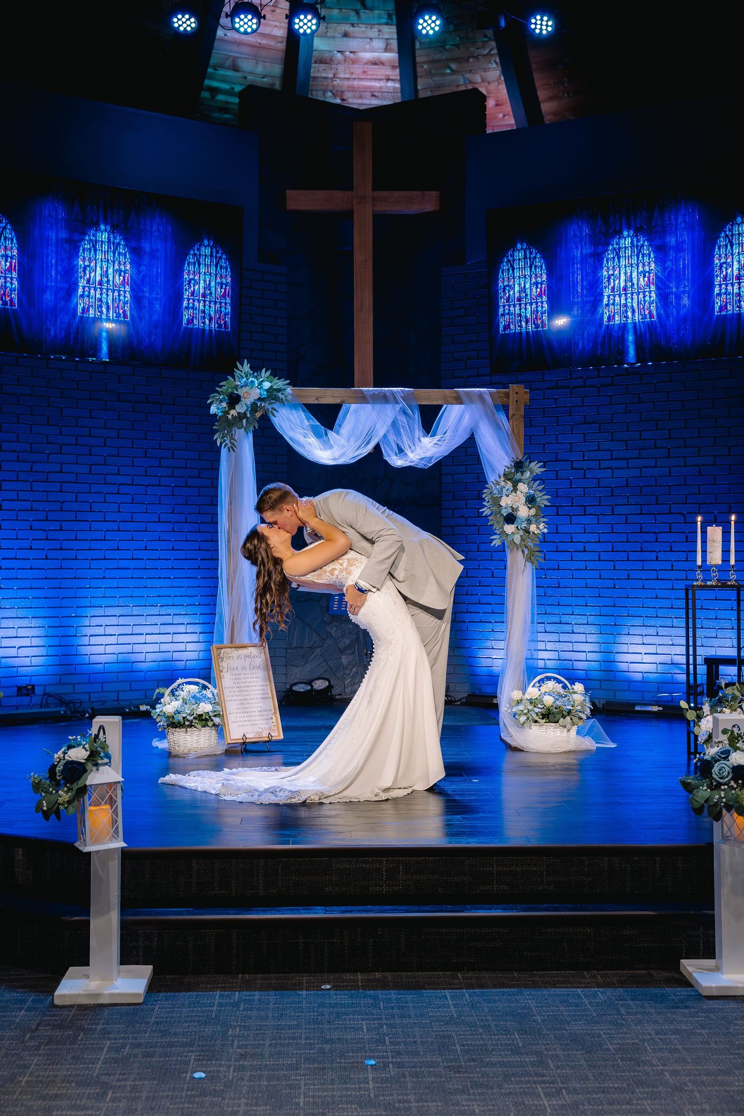 Bride and groom share first kiss under floral arch in church with blue uplighting and wooden cross backdrop.