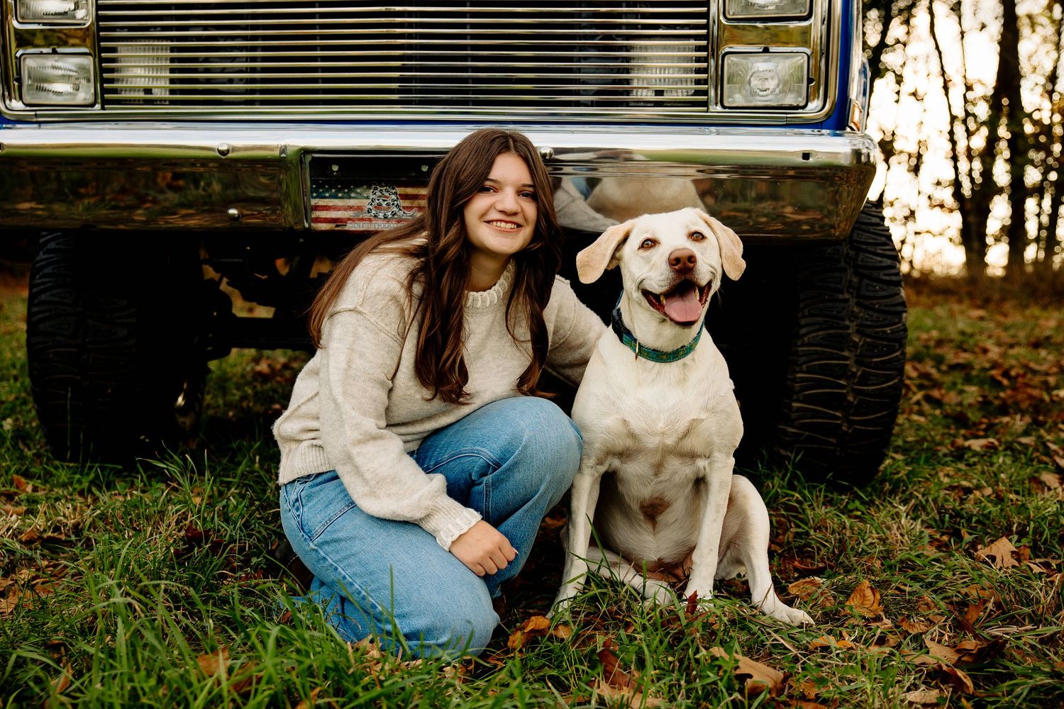 Young woman smiling beside a yellow Labrador Retriever in front of a vintage truck outdoors.