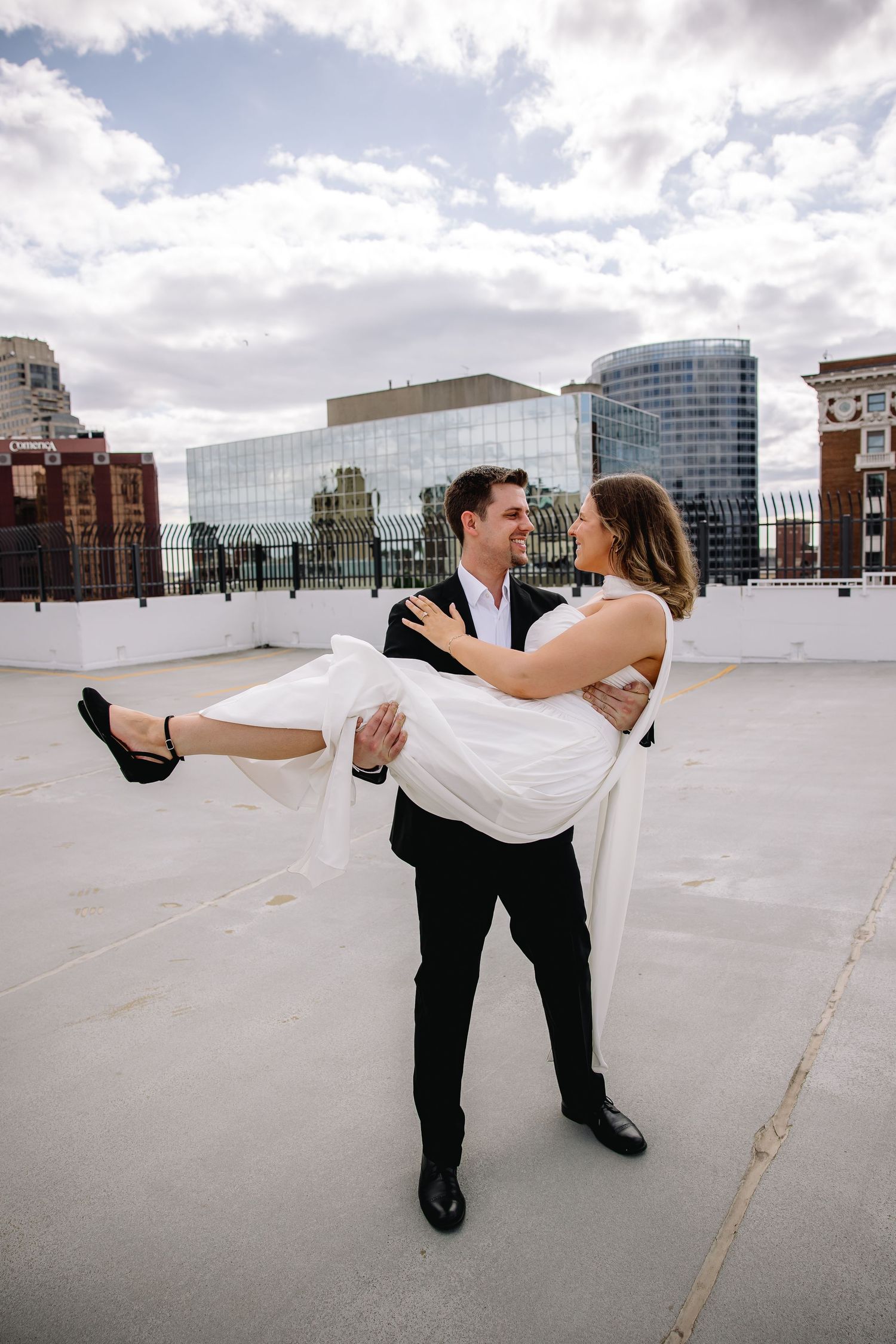 Groom carrying bride in white dress on rooftop with city skyline backdrop on their wedding day.