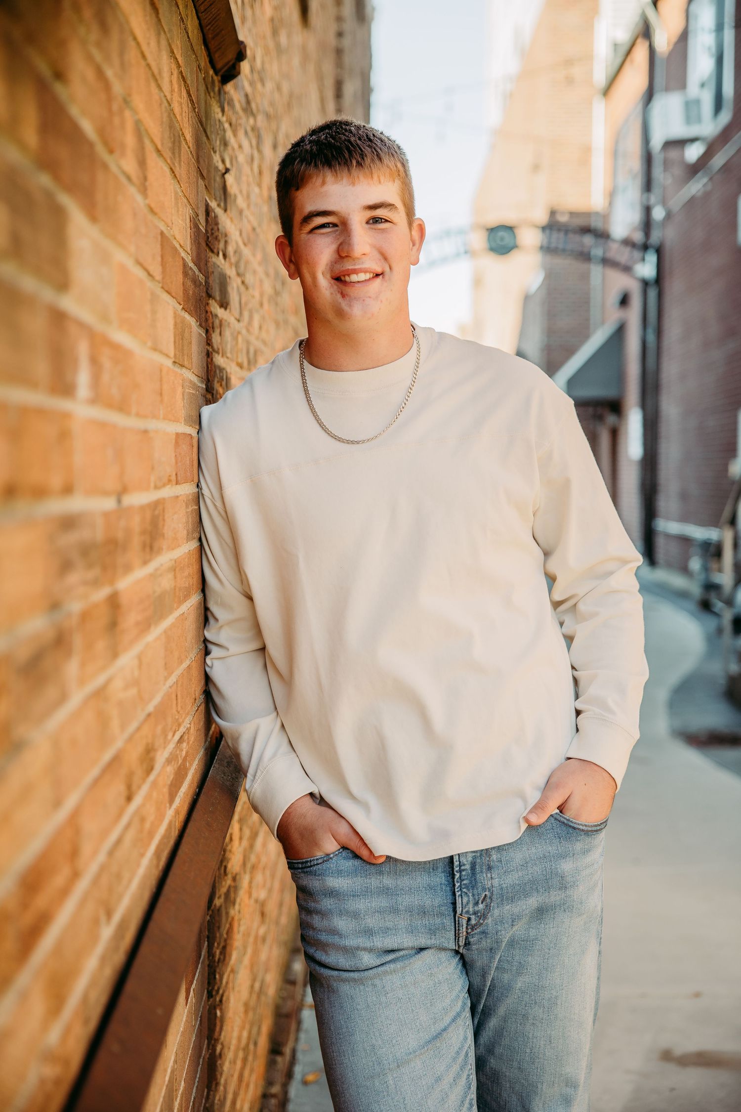Smiling teen boy in cream sweatshirt and jeans leaning against brick wall in urban alley.