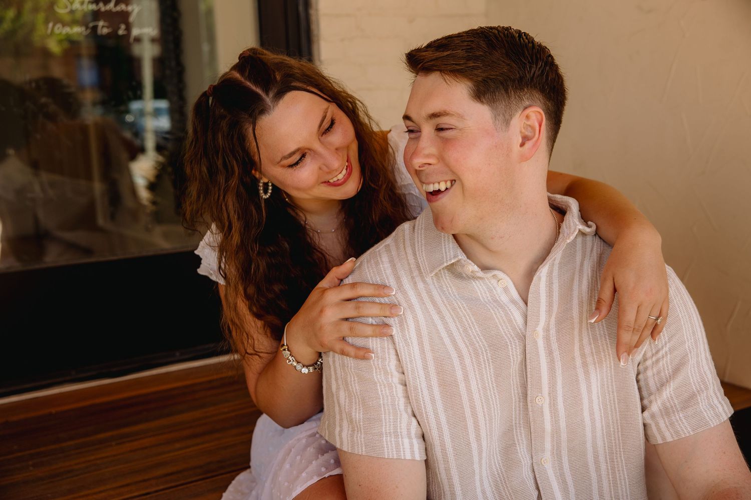 A laughing couple shares a joyful moment, woman in white dress embracing man in striped shirt outdoors.