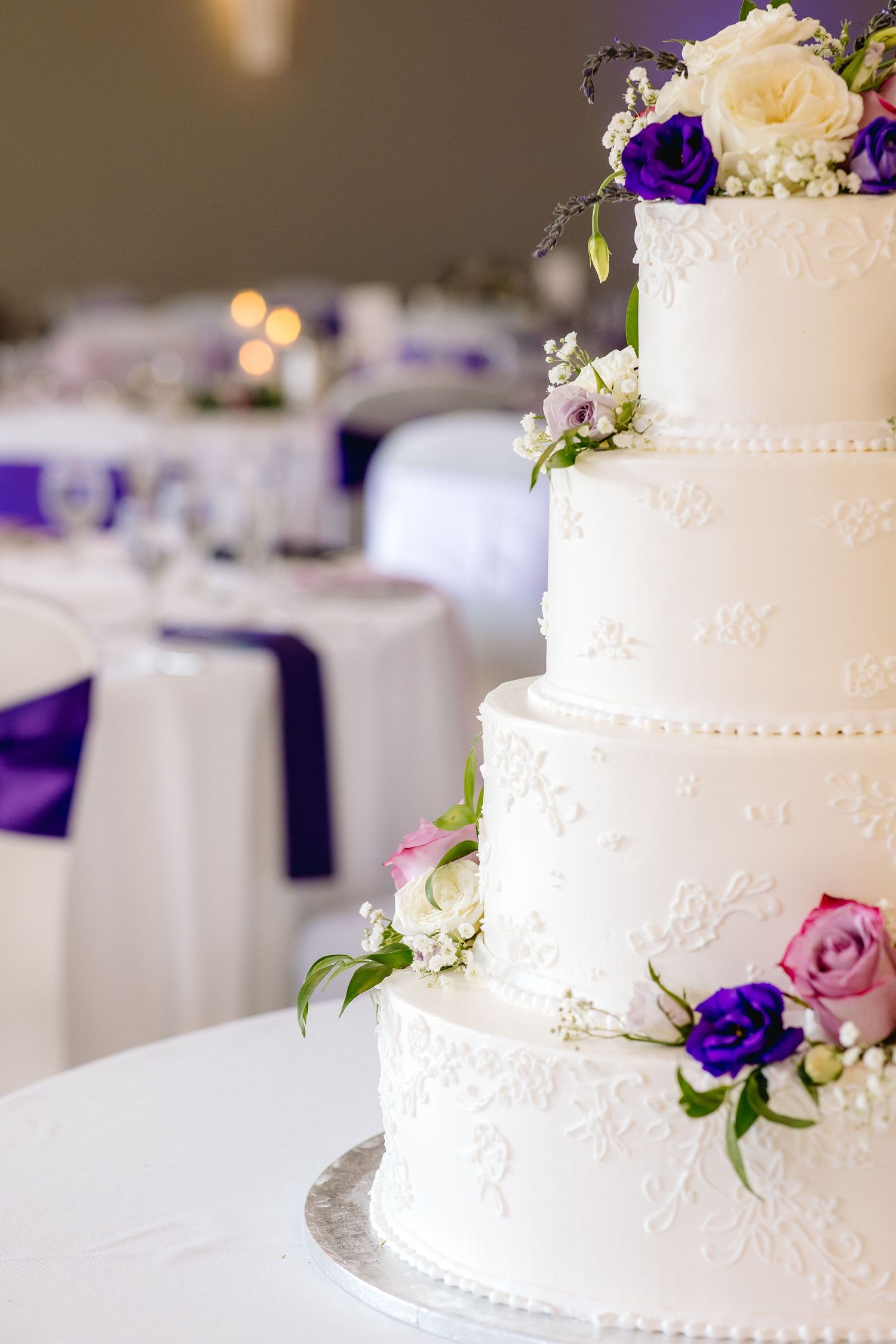 Elegant four-tier white wedding cake adorned with purple and pink flowers, displayed in a decorated reception hall.