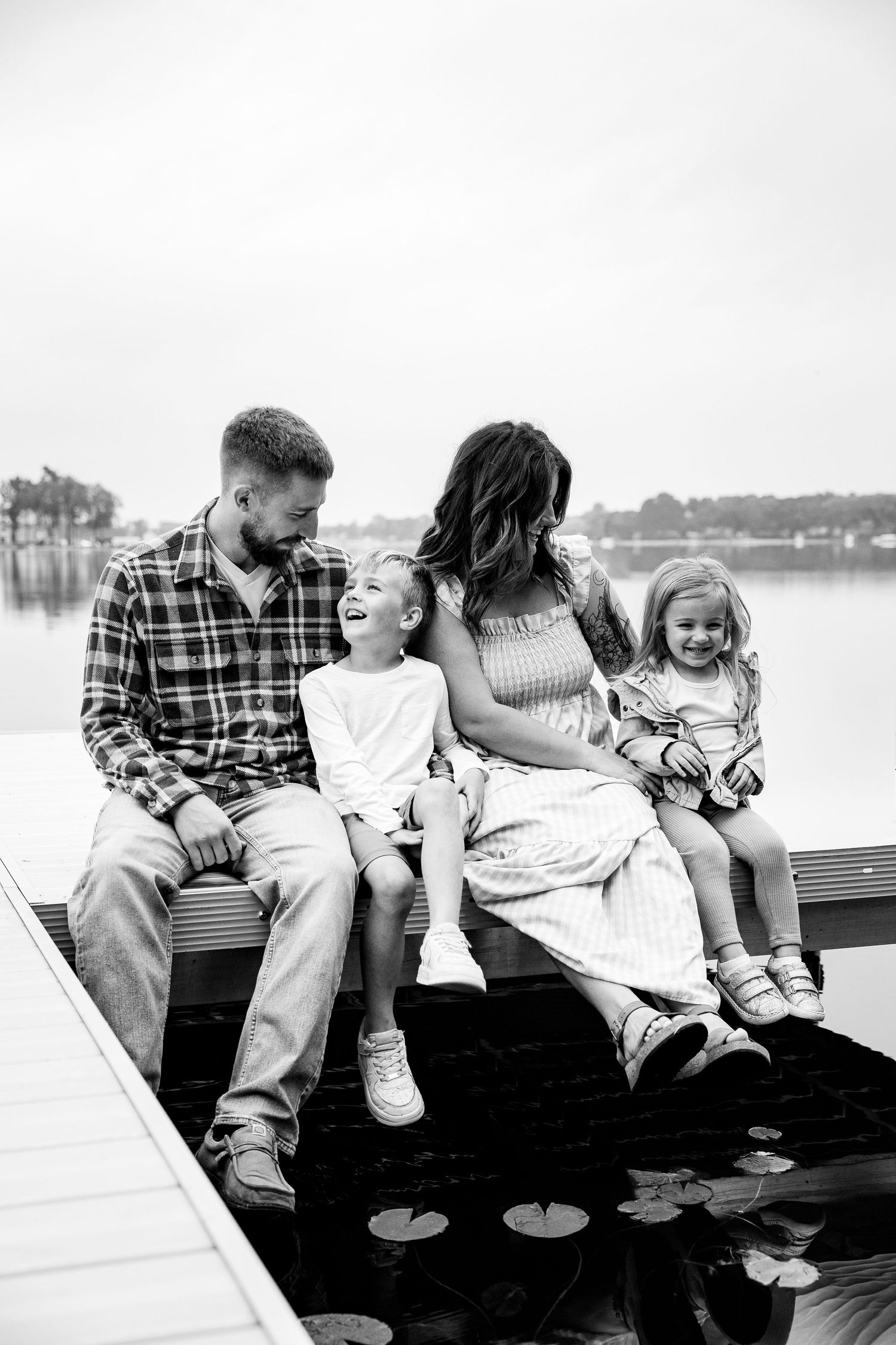 Black and white photo of a happy family of four sitting on a dock by a lake, laughing together.