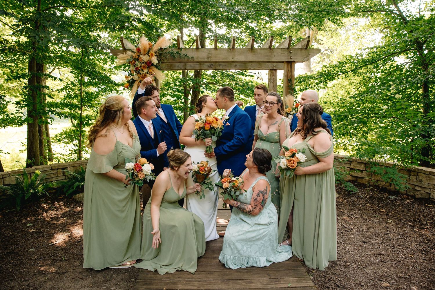 Wedding party in sage green bridesmaid dresses and blue suits gathered under a wooden pergola surrounded by lush green trees.
