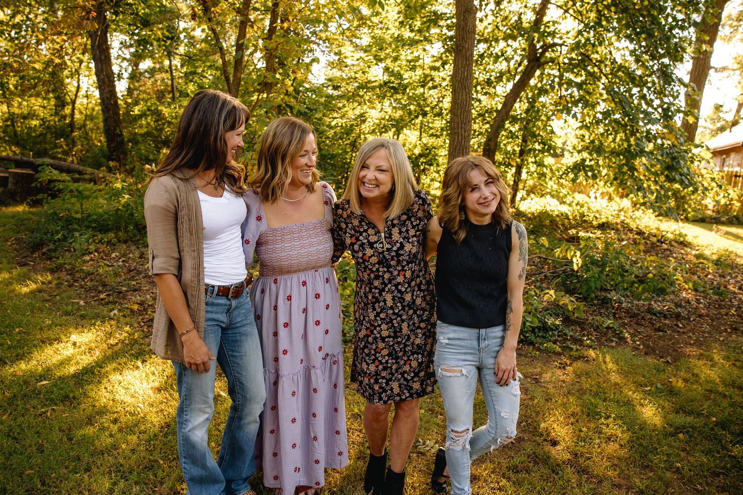 Four women smiling and embracing outdoors in a sunlit garden with lush green trees in the background.