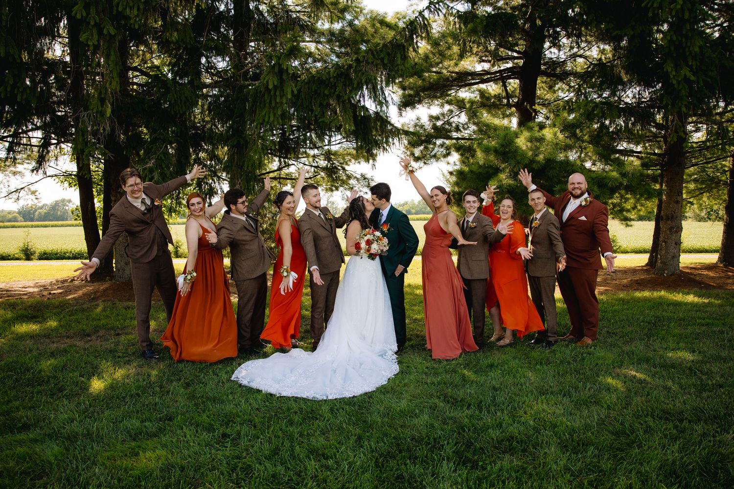 Wedding party in autumn colors, bridesmaids in rust and brown dresses, groomsmen in suits, couple kissing outdoors.