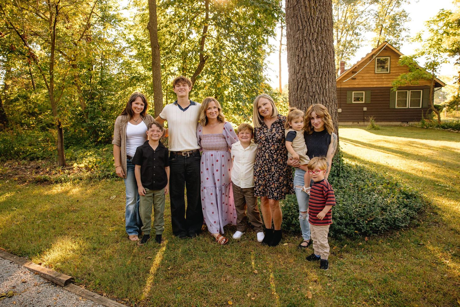 Large family group of adults and children smiling outdoors on green lawn with trees and wooden cabin in background.