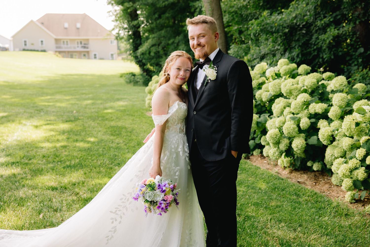 Bride and groom smiling outdoors in wedding attire, bride holding colorful bouquet, green hydrangeas and lawn in background.
