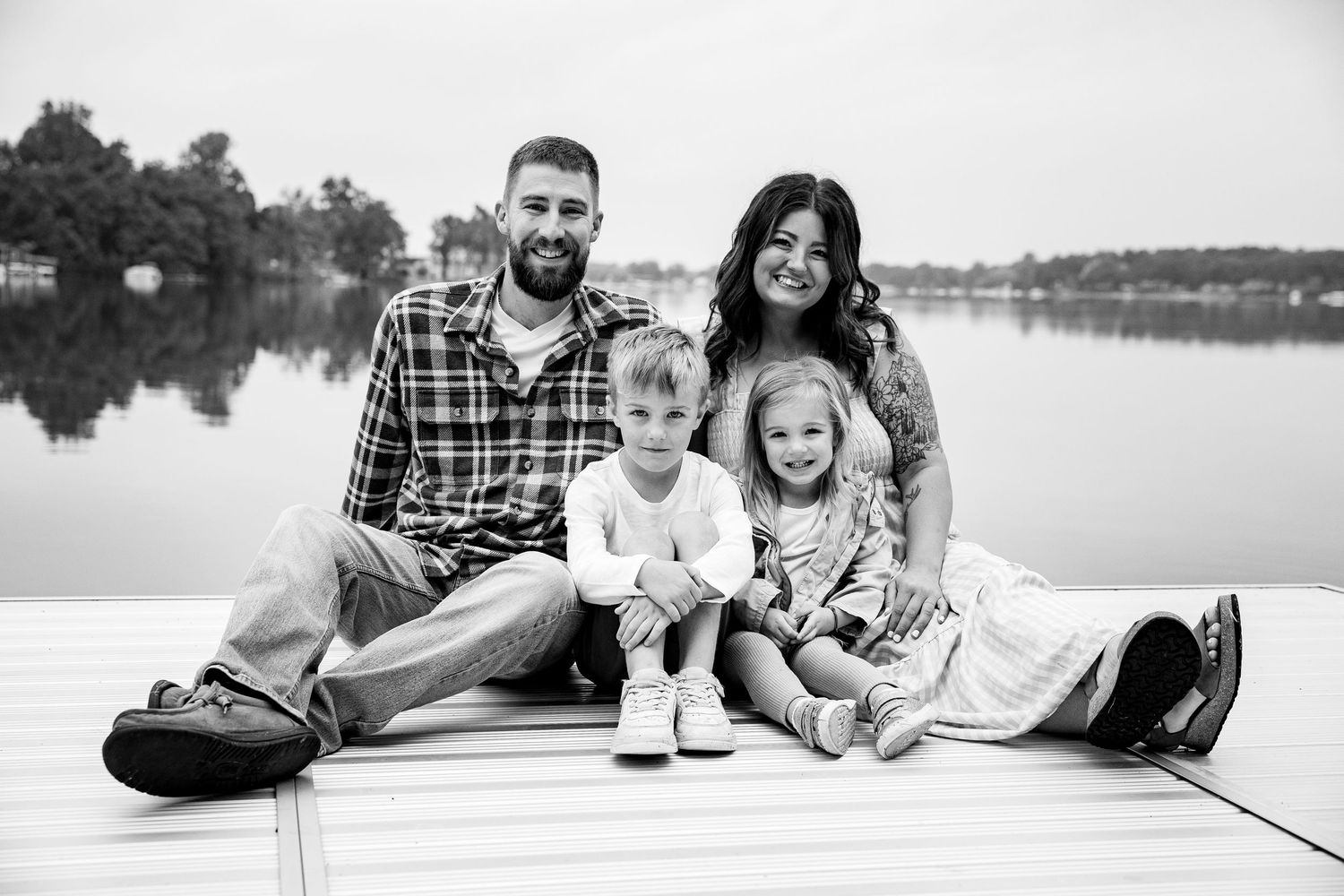 Black and white family photo of parents with two young children sitting on a dock by a lake.