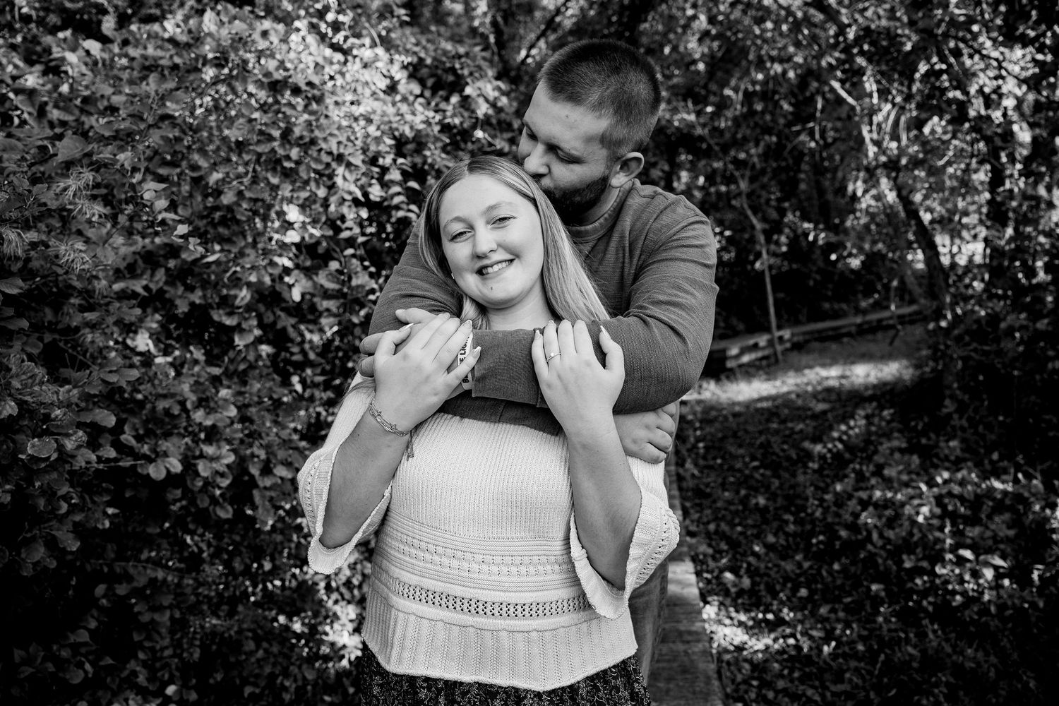 Black and white photo of a couple embracing outdoors, man hugging woman from behind among lush green trees.