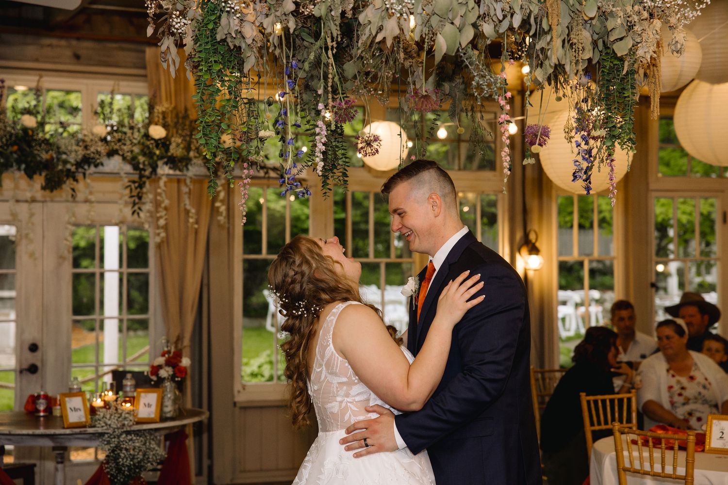 Bride and groom share first dance under floral ceiling with hanging lights at elegant indoor wedding reception.