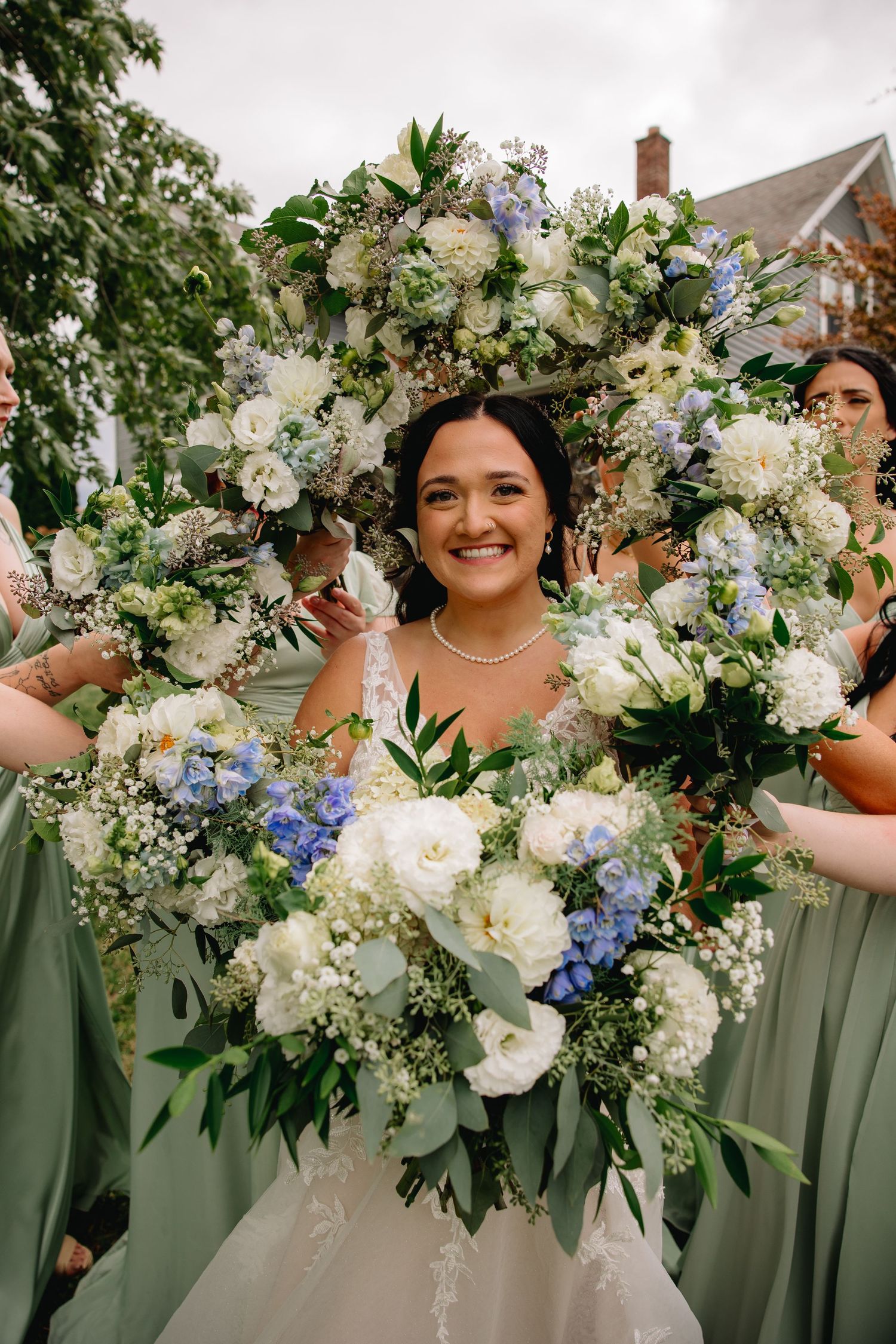 Smiling bride surrounded by bridesmaids holding lush white and blue floral bouquets at an outdoor wedding ceremony.