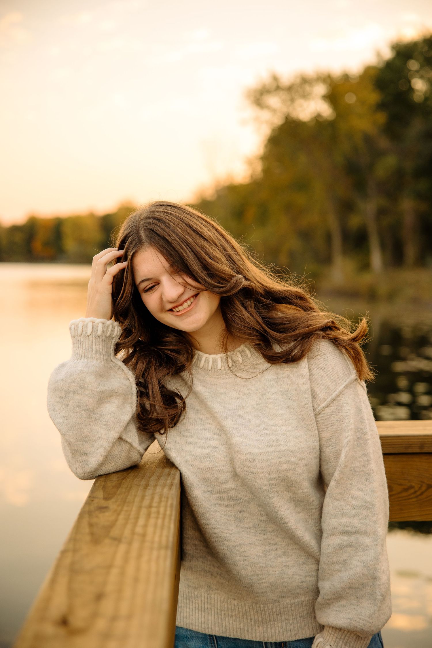 Young woman in beige sweater smiling on wooden dock by lake at golden sunset with autumn trees.