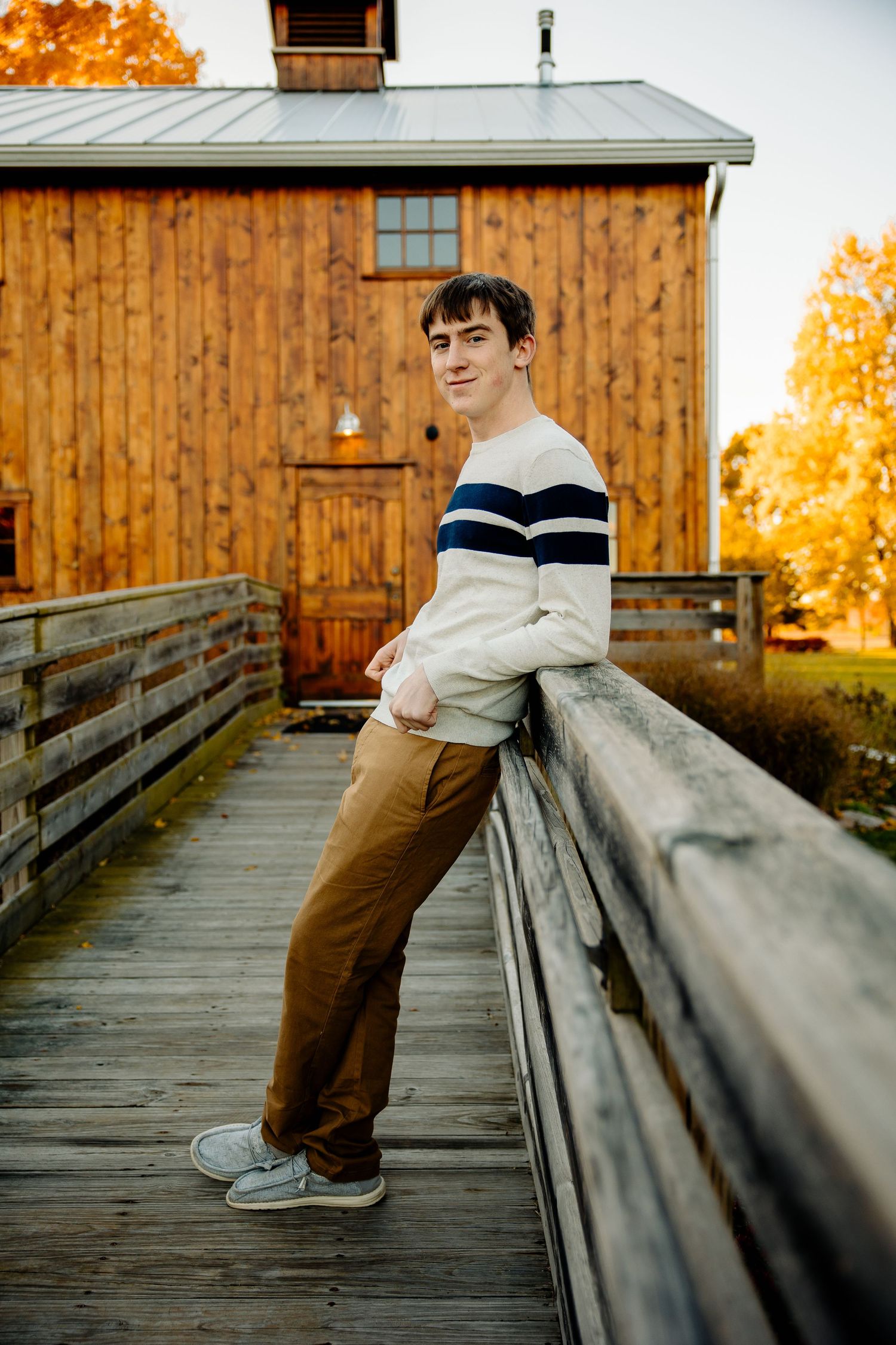 Young man in striped sweater and tan pants leaning on wooden bridge railing near rustic barn in autumn.