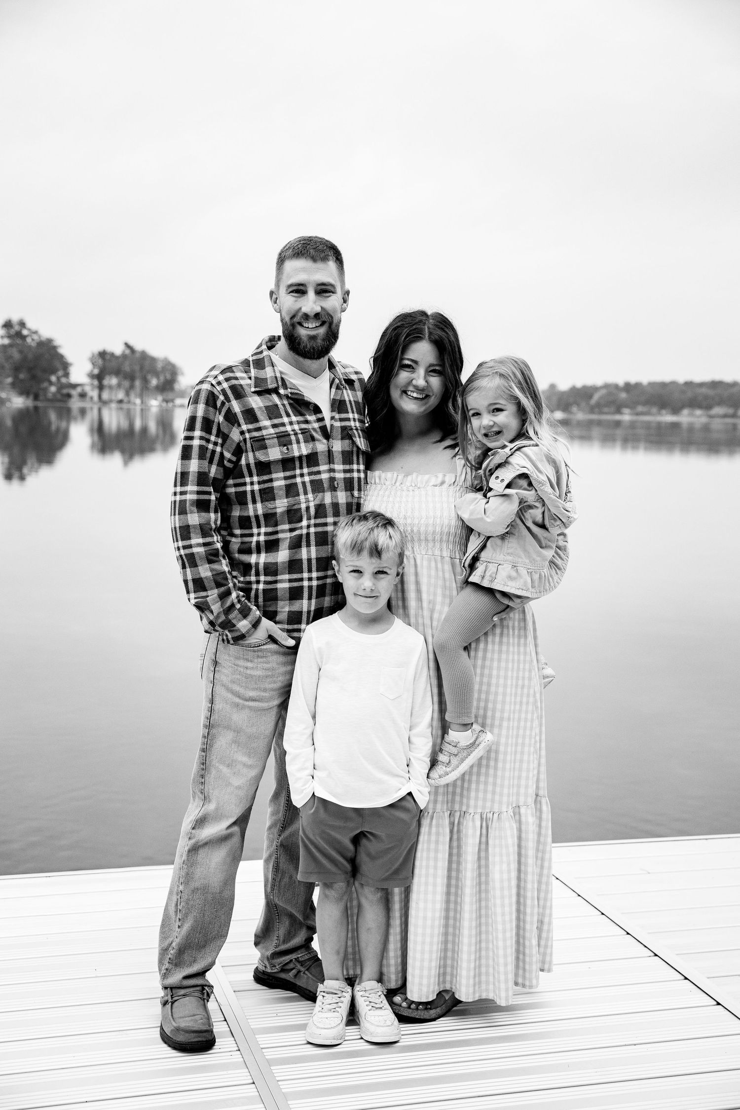 Black and white family portrait of parents and two young children standing on a dock by a calm lake.