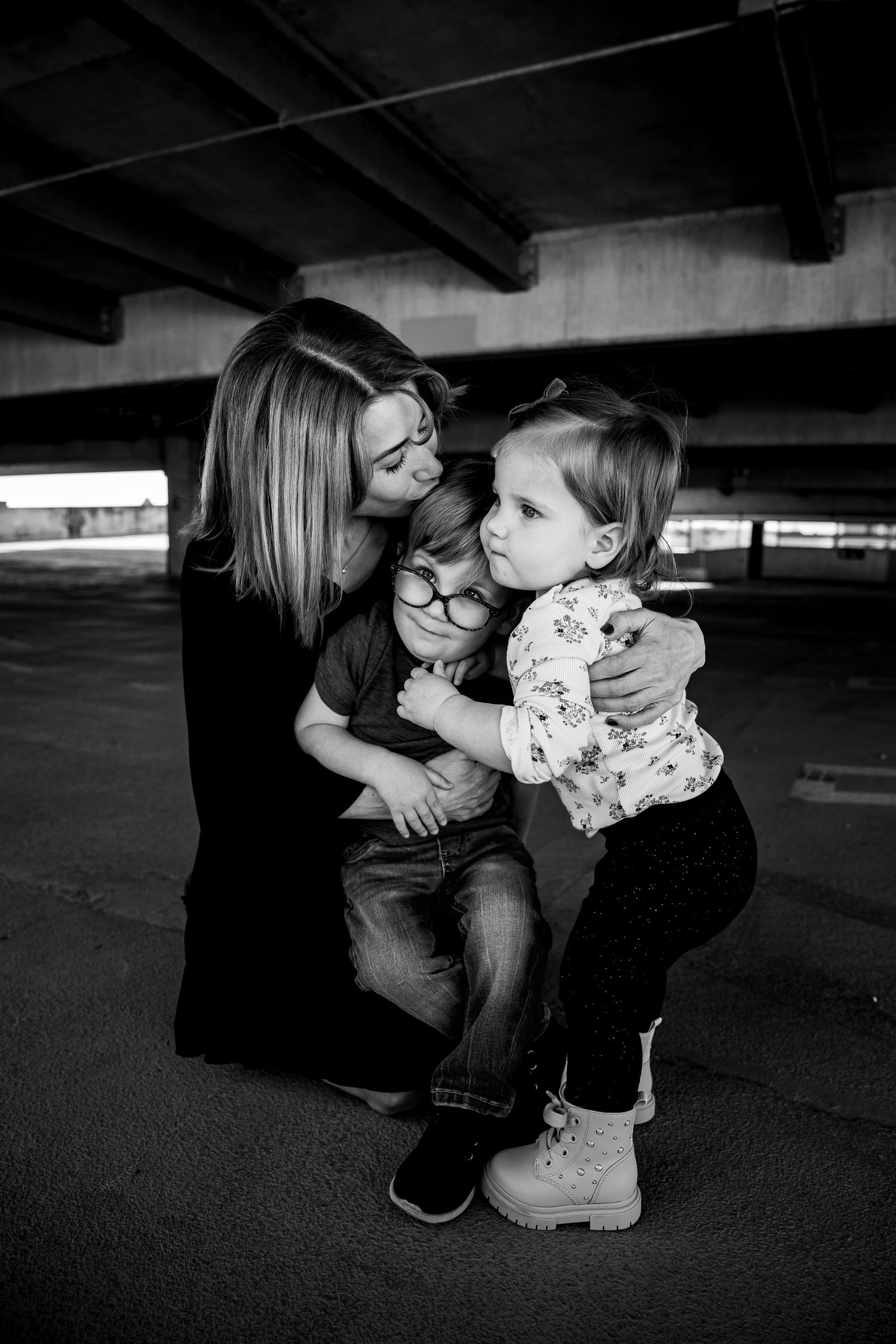 Black and white photo of a mother hugging her two young children in an urban parking garage setting.