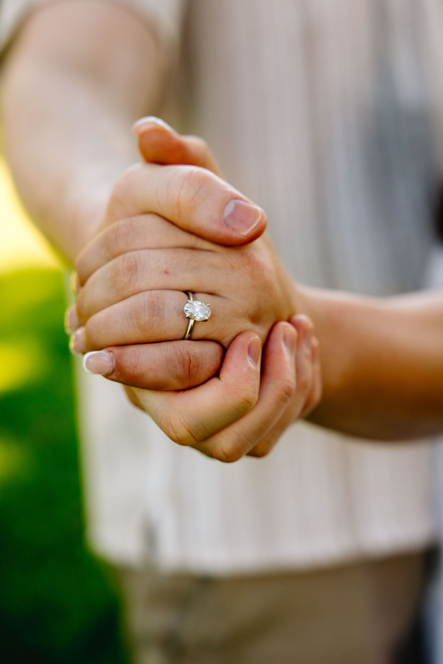 Two hands clasped together showing a beautiful solitaire diamond engagement ring on a gold band outdoors.