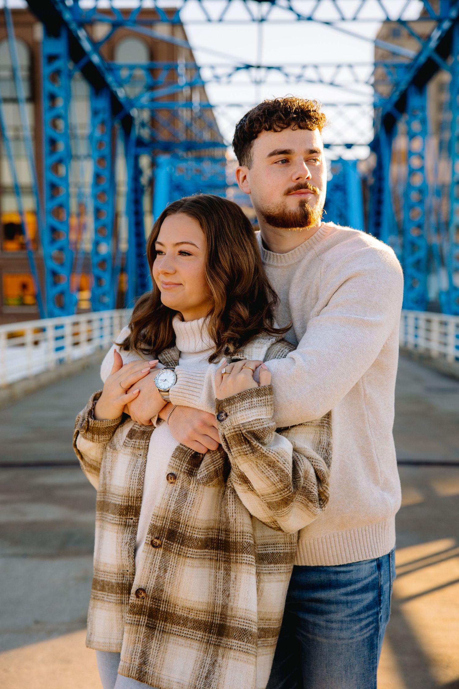 Couple embracing on a blue bridge, woman in plaid jacket with watch, man in cream sweater, sunny day.