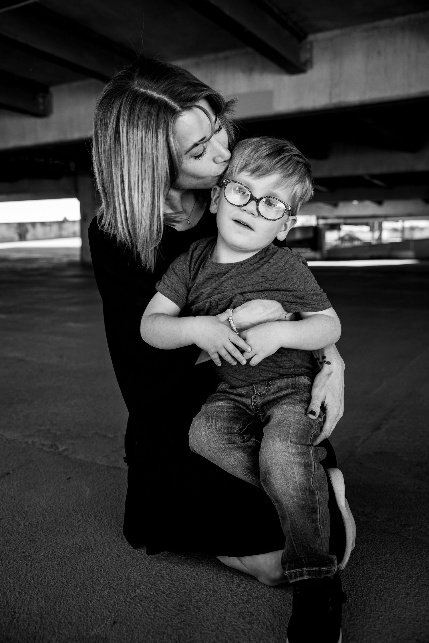 Black and white photo of a mother kissing her young son wearing glasses as he sits on her lap under a bridge.