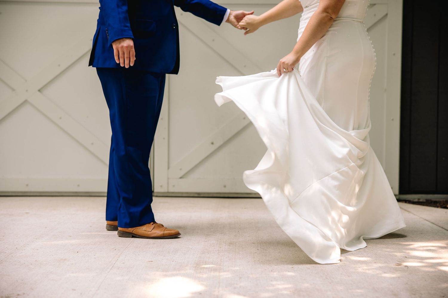 Bride in white gown twirling while groom in blue suit holds her hand outside barn doors.