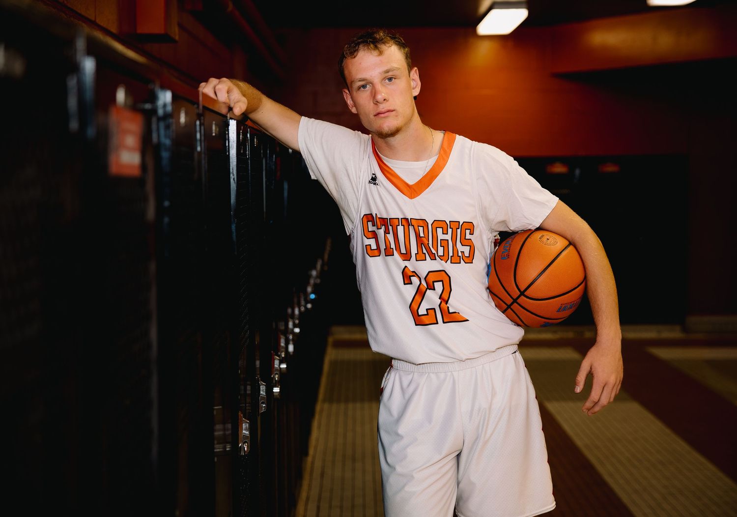 Sturgis basketball player #22 poses with ball in locker room wearing white and orange uniform.