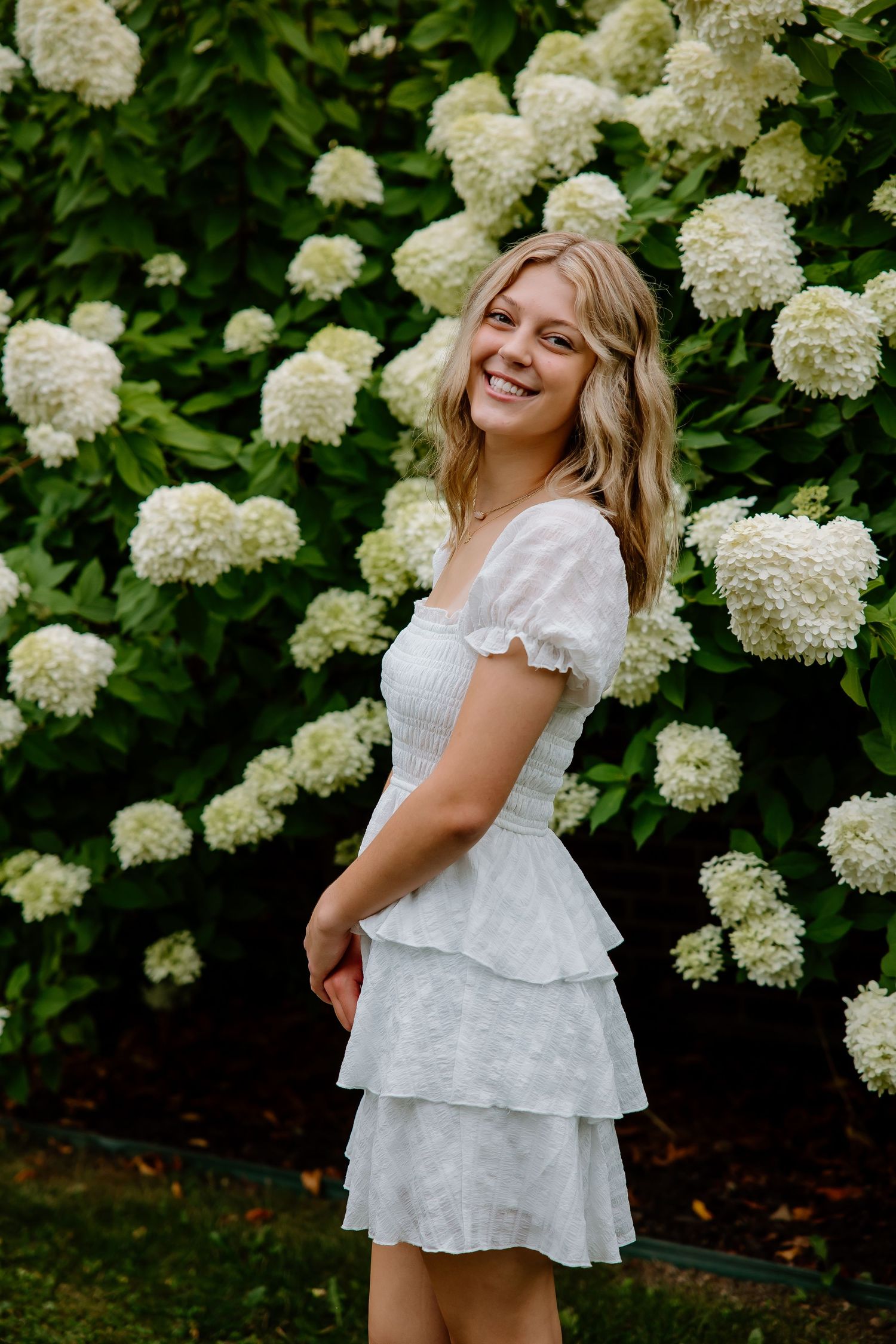 Young blonde woman in white tiered dress smiling in front of blooming white hydrangea bushes outdoors.