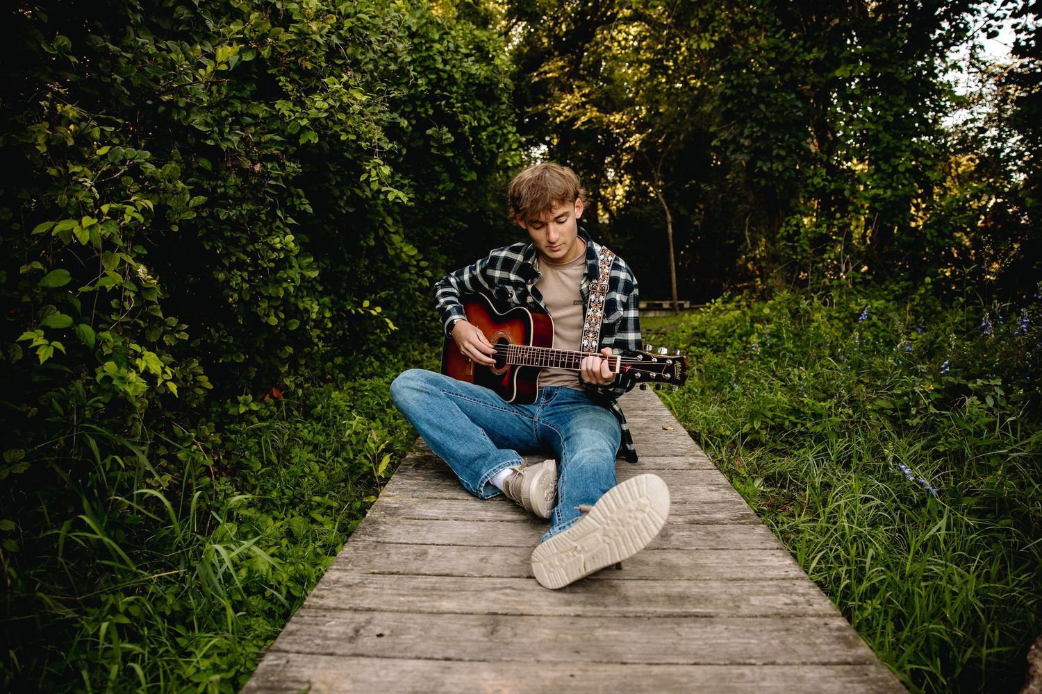 Young man playing acoustic guitar while sitting cross-legged on a wooden path surrounded by lush green trees.