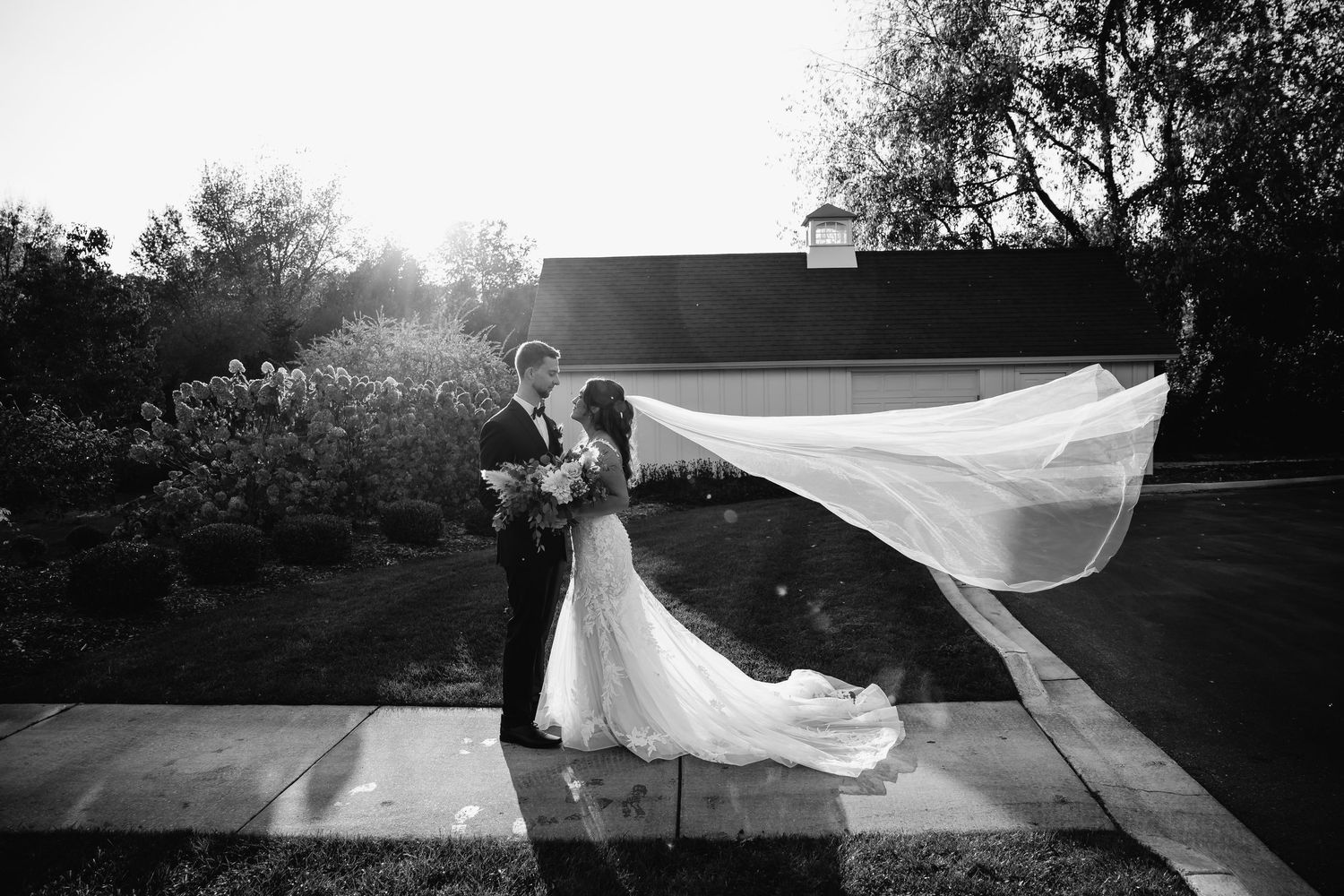 Black and white wedding photo of bride and groom kissing as veil flows dramatically in the wind outdoors.