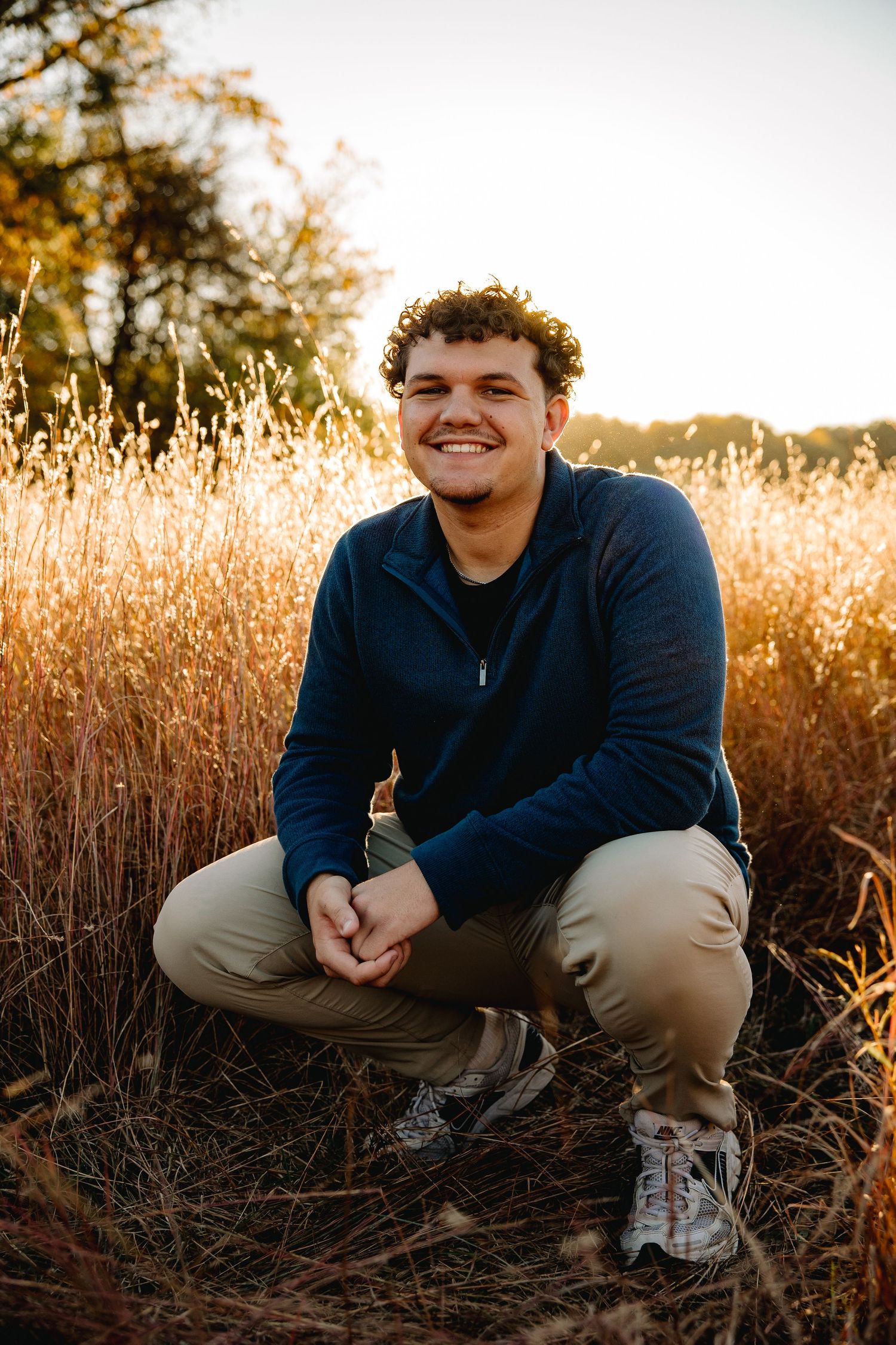 Young man with curly hair crouching in golden field at sunset, smiling, wearing blue jacket and khaki pants.