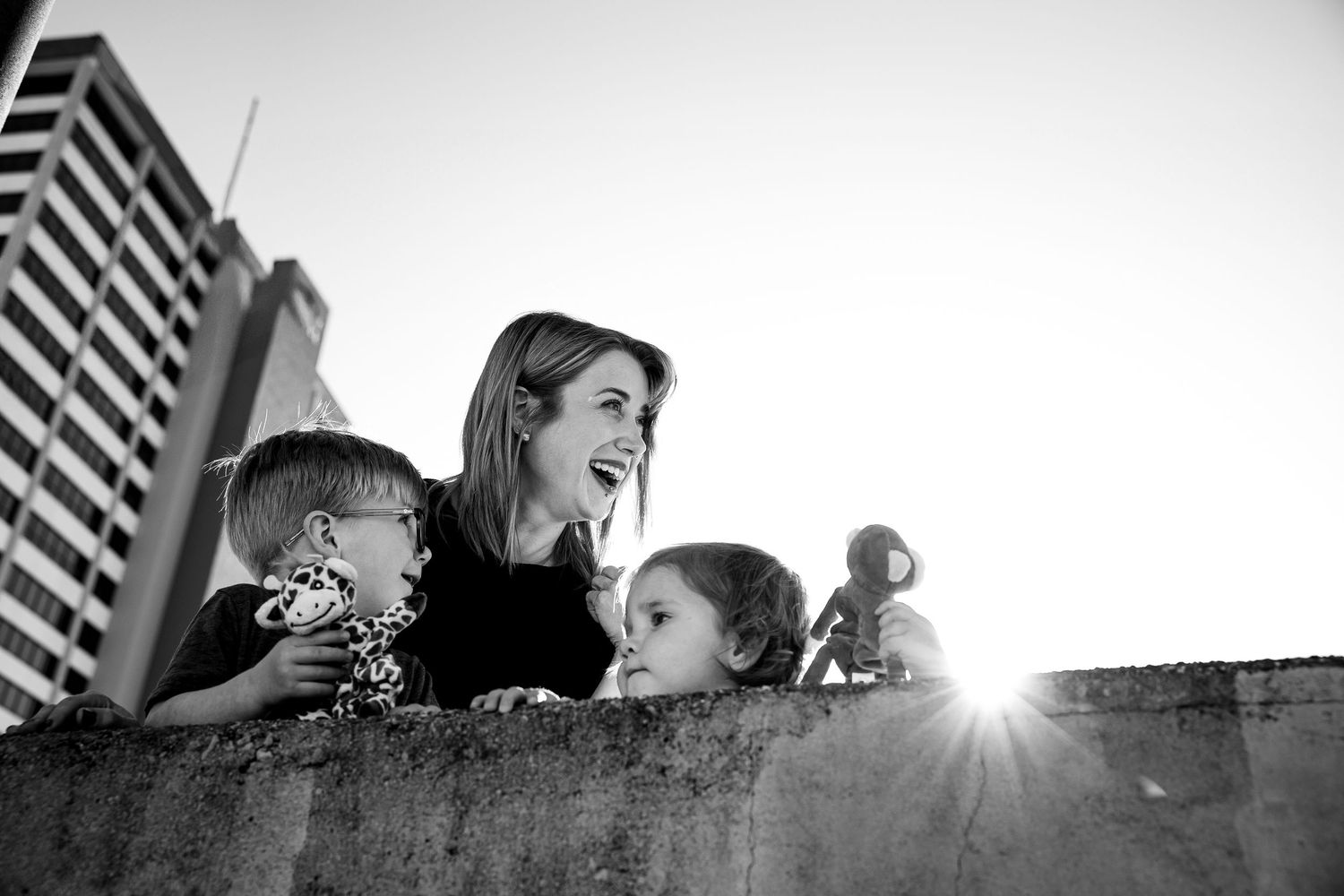 Black and white photo of a laughing woman and two children peering over a concrete wall with city buildings in background.