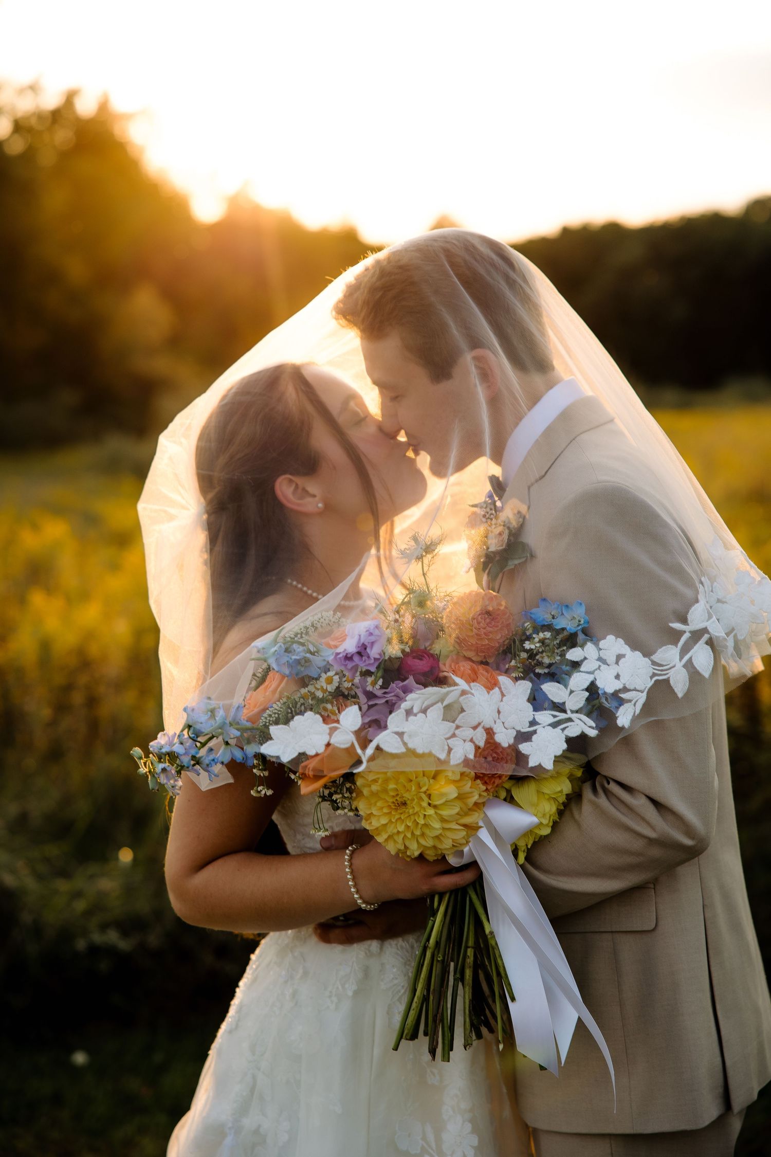 Bride and groom kissing under veil at golden hour, holding colorful wildflower bouquet in sunny outdoor field.