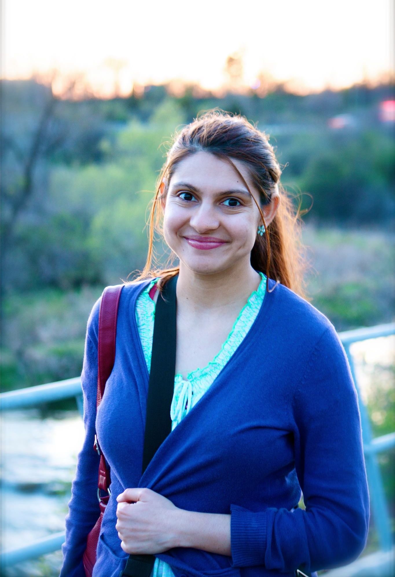 A person in a navy blue cardigan with a backpack stands outdoors against a blurred natural background during sunset.