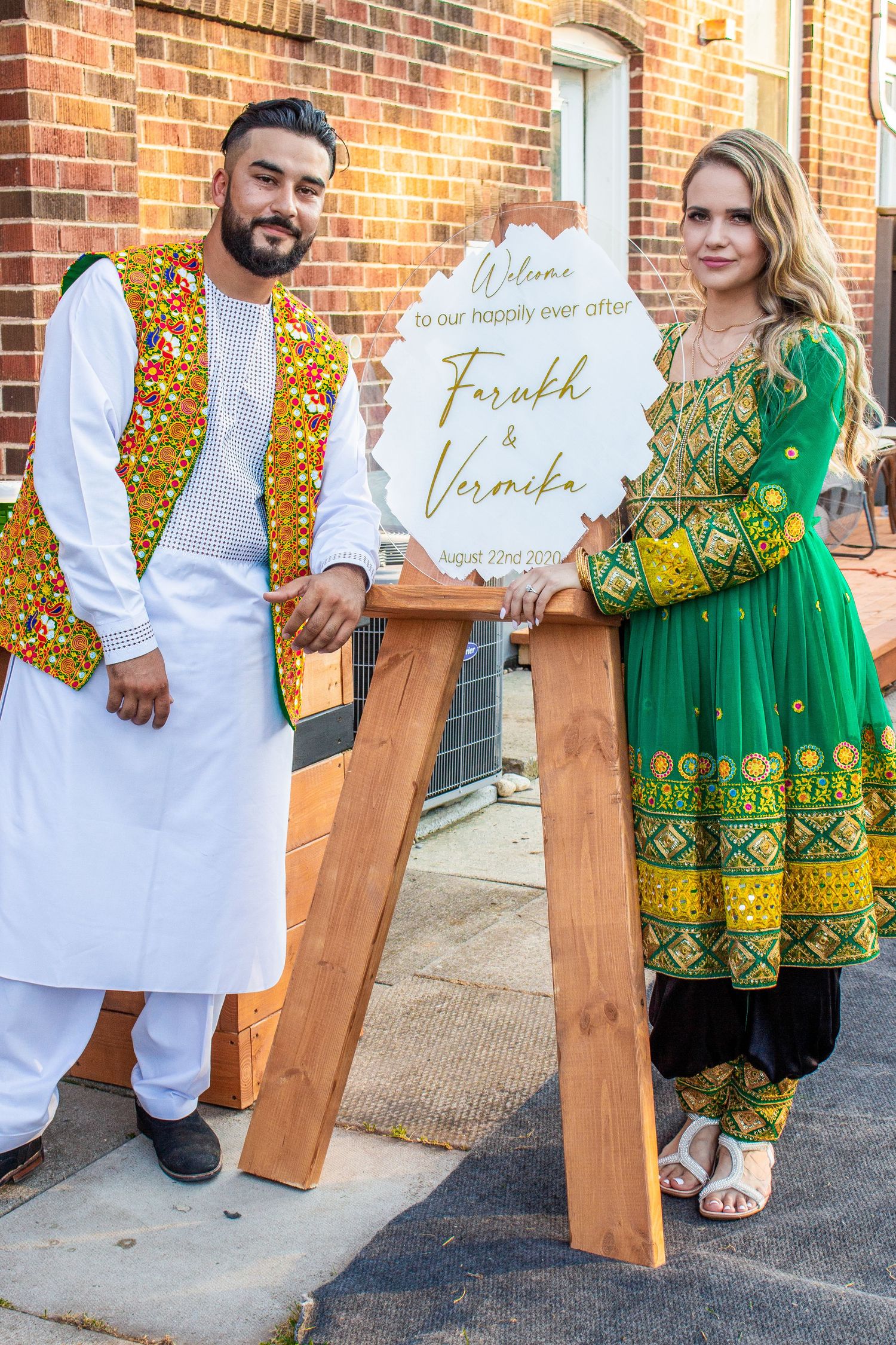 Toronto Event Photographer - Couple standing beside wooden welcome sign wearing colorful traditional Pakistani wedding outfits.